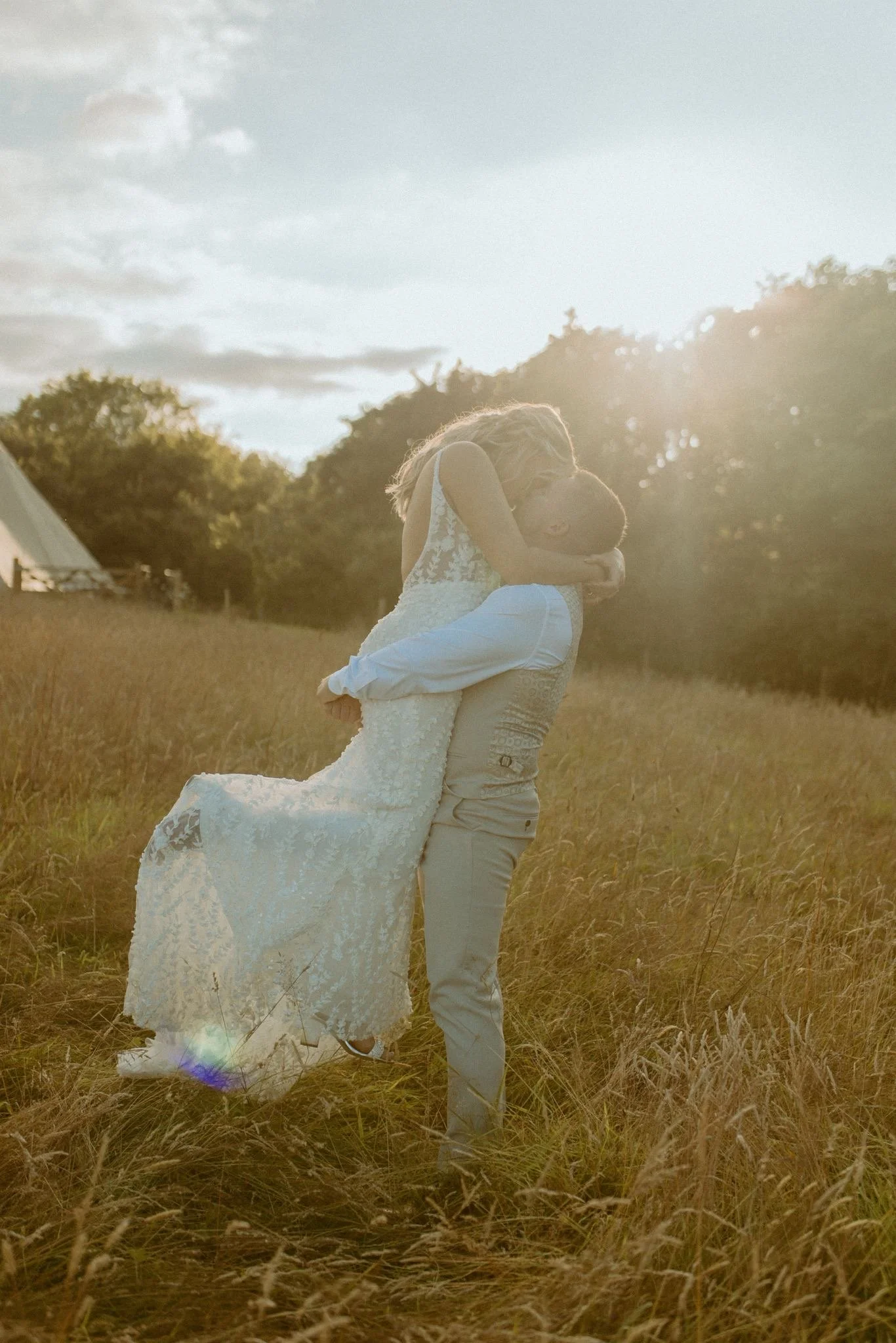 A couple, with the man lifting the woman in a white lace wedding dress, embraces in a grassy field at sunset.