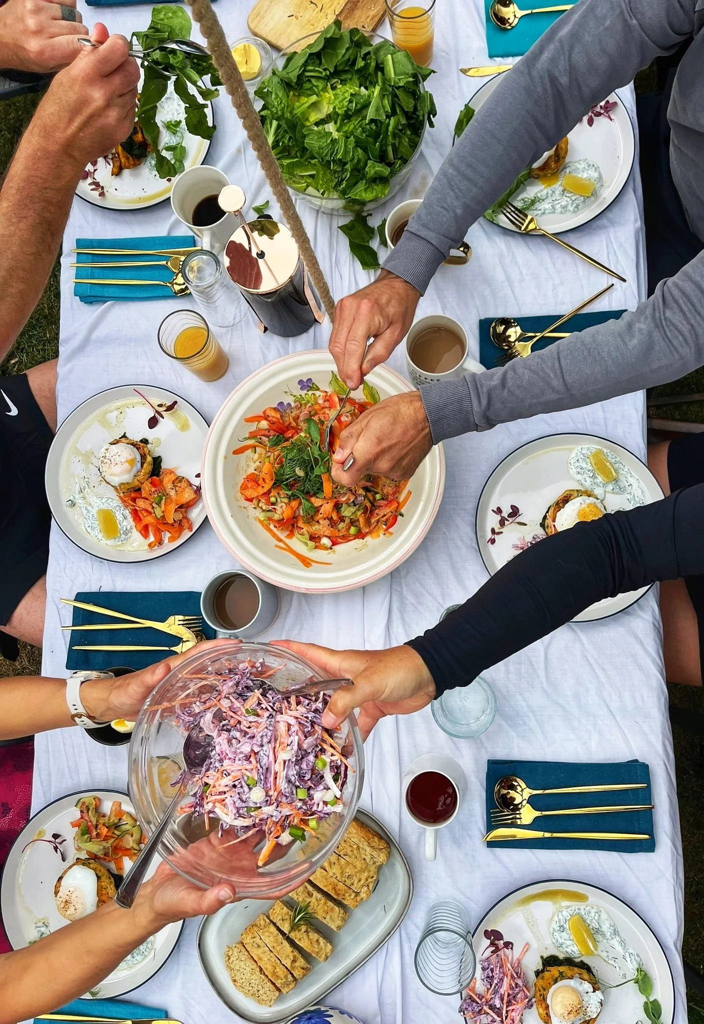 A family gathering with a white tablecloth, serving dishes, salads, bread, and drinks, as people pass bowls and prepare to eat.
