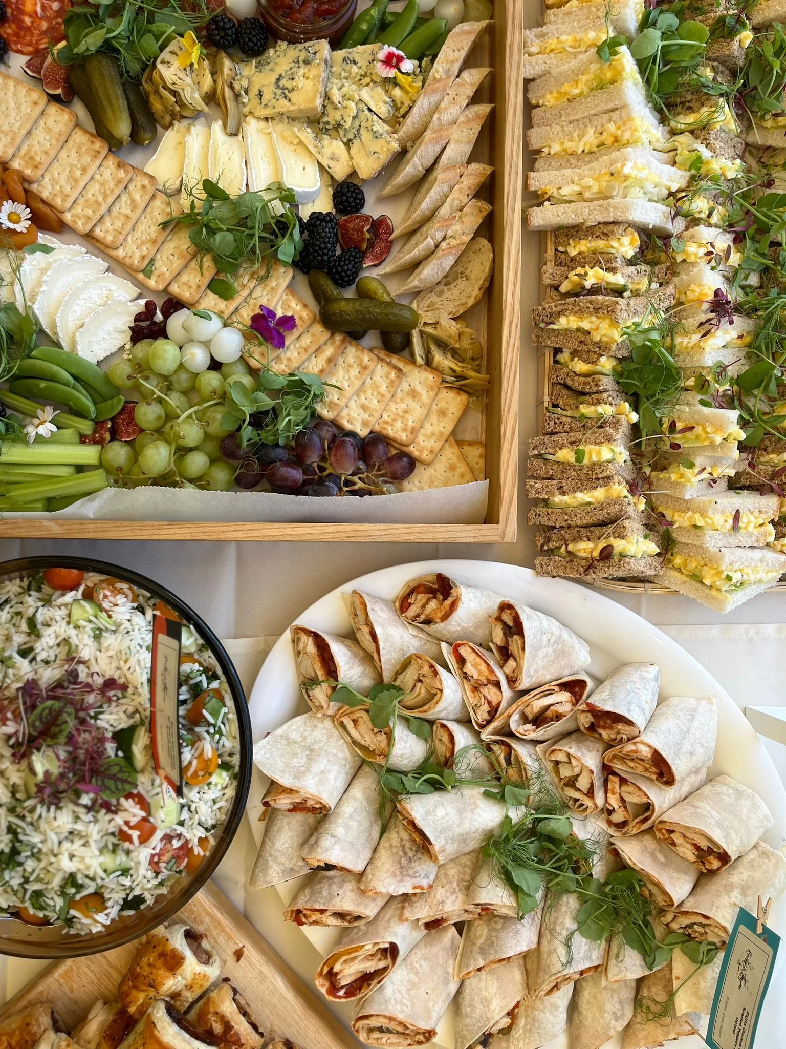 Assorted cheese platter with grapes, crackers, fresh herbs, and berries; finger sandwiches with egg salad and herbs; sliced wrap sandwiches with vegetables and meat; chopped vegetable and rice salad.