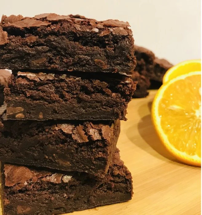 A stack of four chocolate brownies next to a sliced orange on a wooden surface.