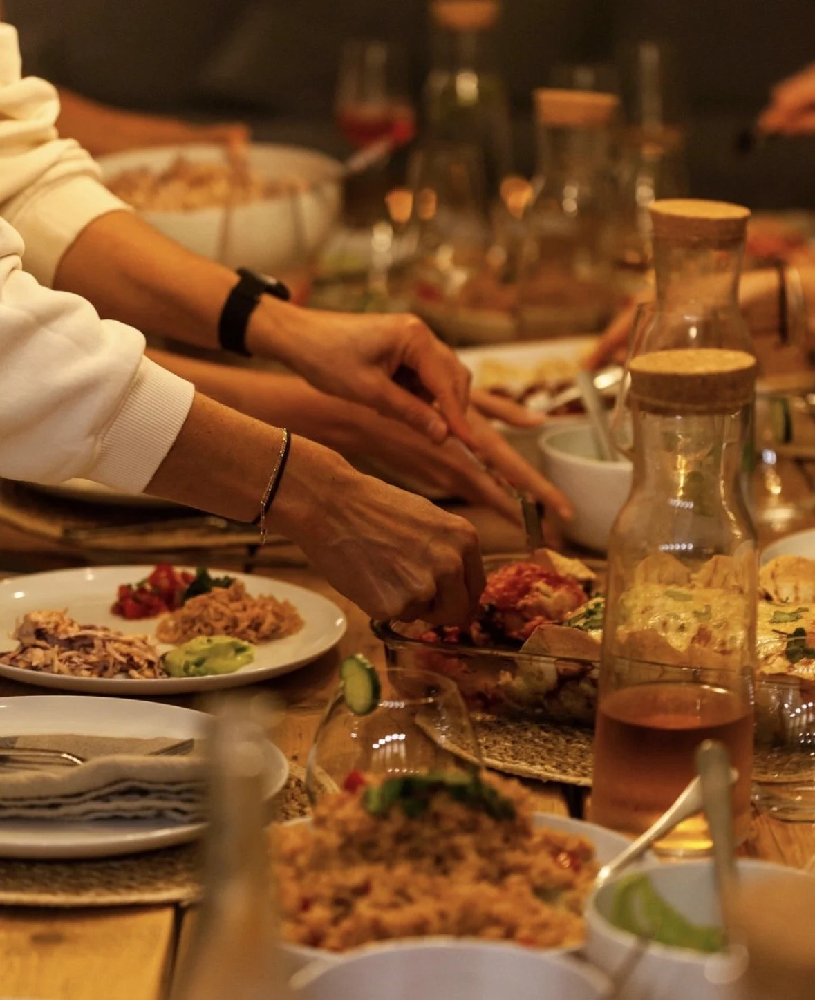 People serving themselves food at a gathering with plates, bowls, and drinks on a wooden table.