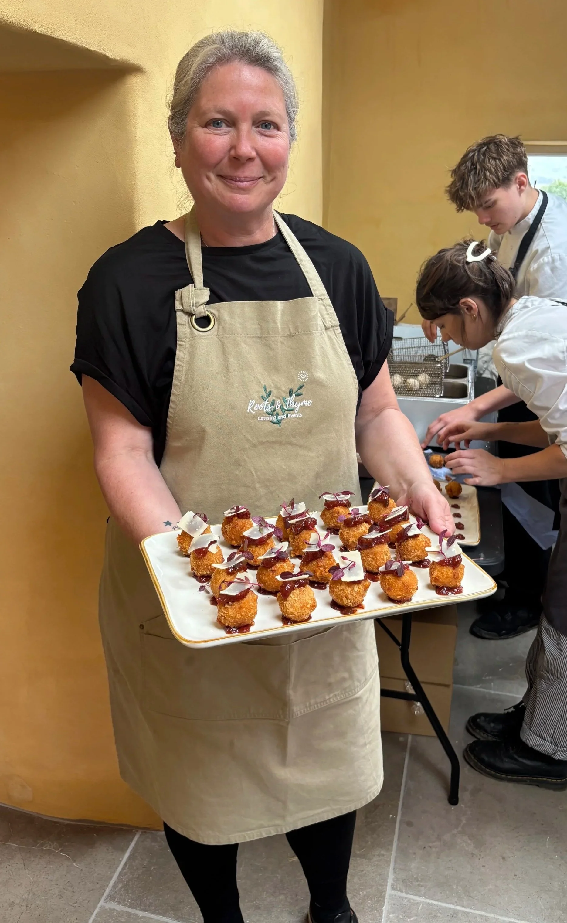 A woman wearing a beige apron holding a tray of bite-sized appetizers with fried interior, decorated with dollops of sauce and edible flowers, in a kitchen or catering setting with two young chefs in the background.