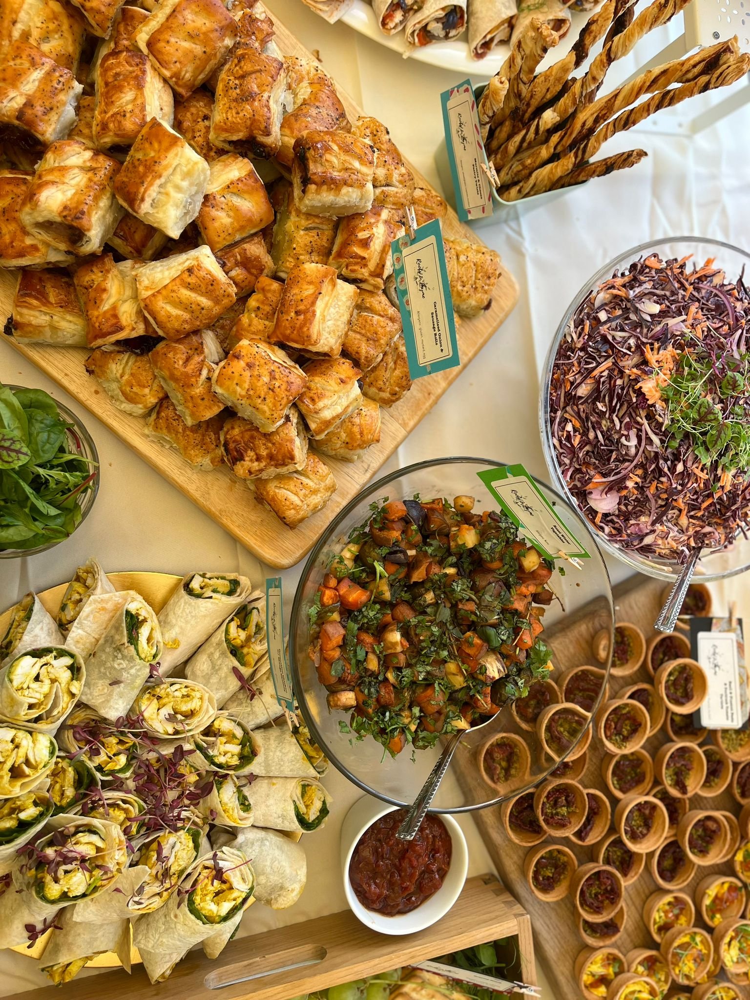 A buffet spread featuring various appetizers including puff pastry bites, veggie wraps, a mixed vegetable salad, coleslaw, and mini taco cups, with some chips and dip, arranged on a white tablecloth.