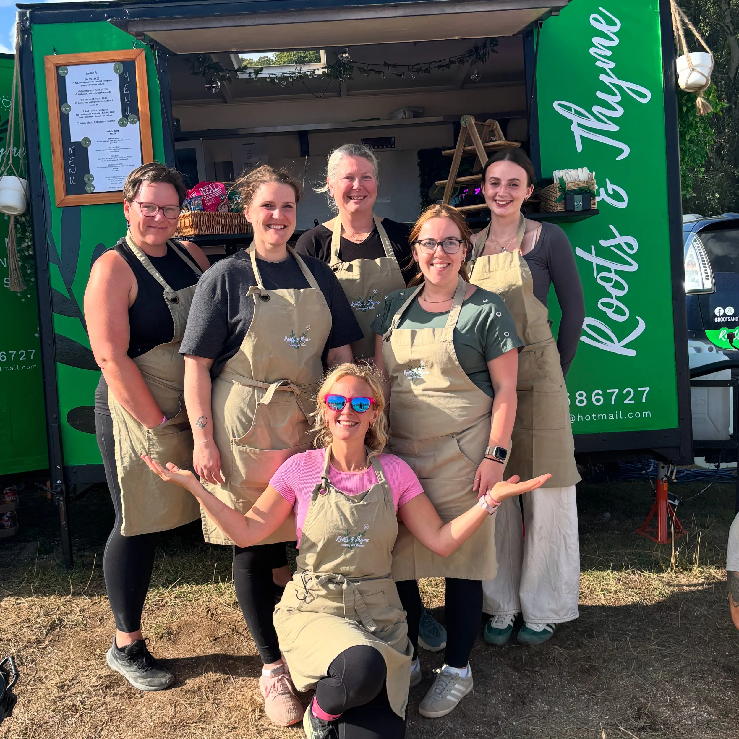 Group of seven women posing in front of a green food truck with the words "Root & Thyme" on it. They are all wearing aprons and smiling, with one woman kneeling in front wearing pink sunglasses and a pink shirt.