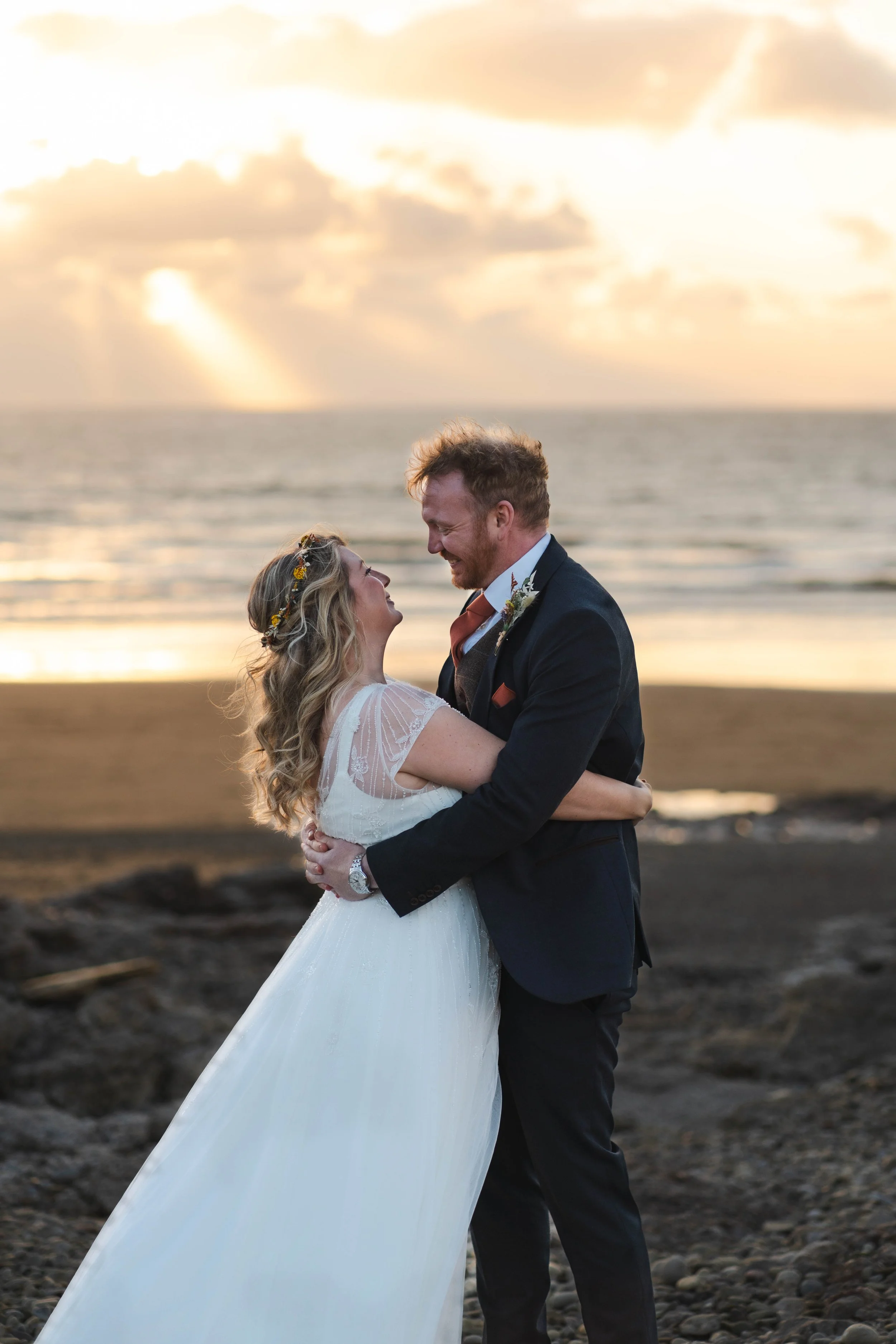A bride and groom embrace on a beach during sunset, smiling at each other.
