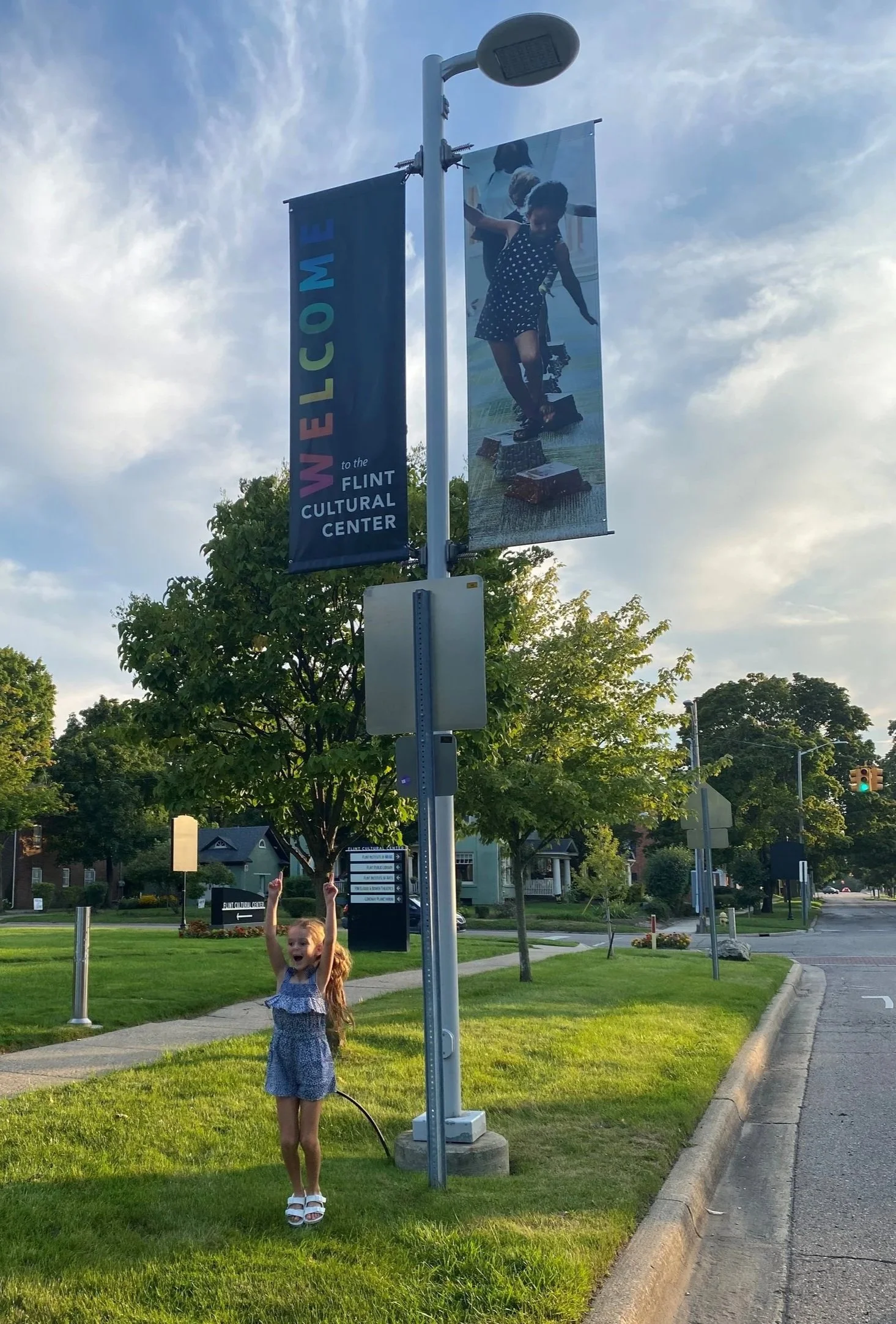 A young girl with long red hair, wearing a blue dress and white sandals, standing on a grassy area near a sidewalk. She is smiling and raising her arms in the air with excitement. Behind her is a park with trees and a street with houses and traffic l