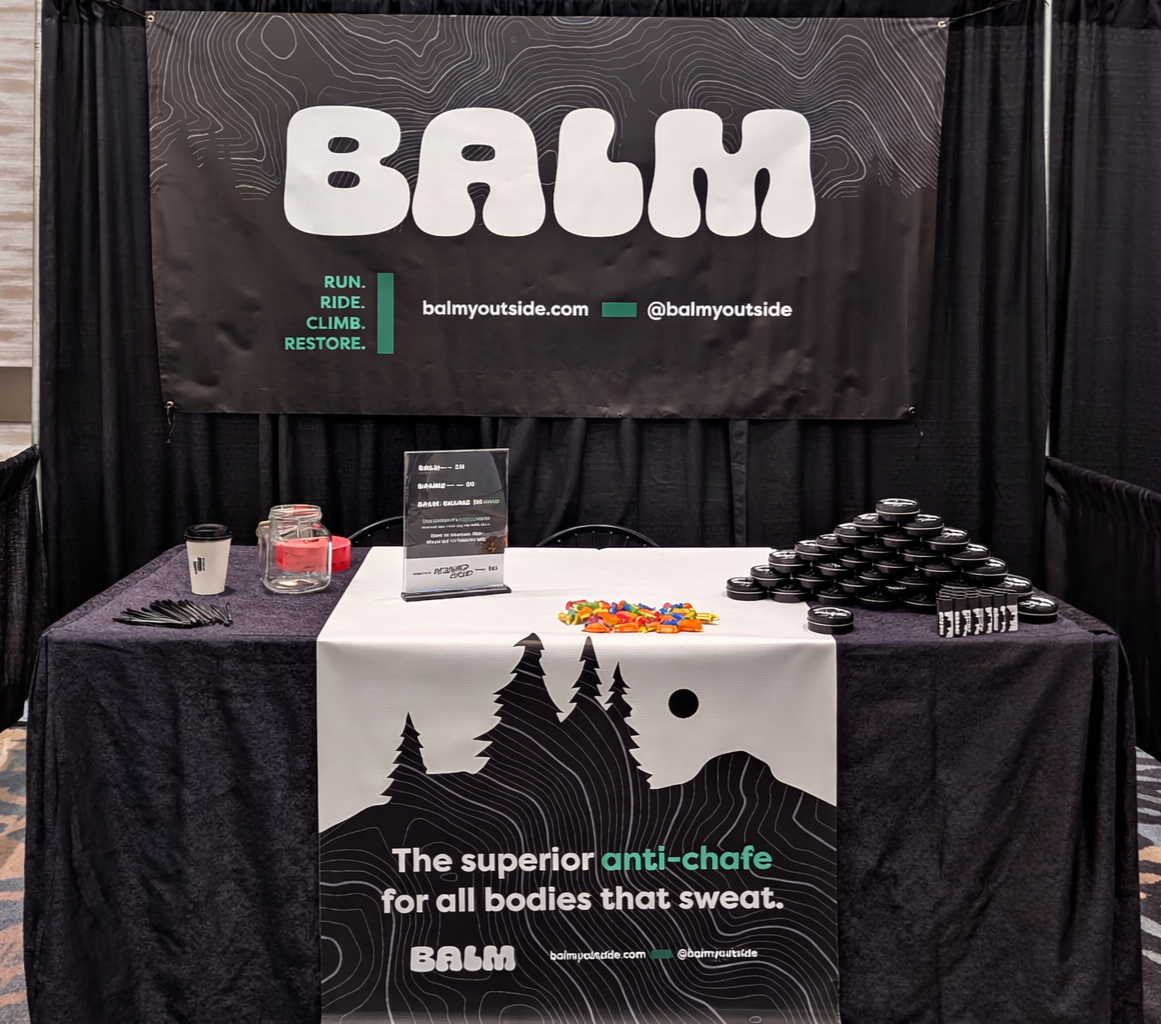 A promotional display for BALM featuring a large black backdrop with white and teal text, a table with black tablecloth, and a white banner with tree silhouette and product information. The table has small containers, a jar with colorful candies, a coffee cup, and a clear acrylic stand with informational paper.