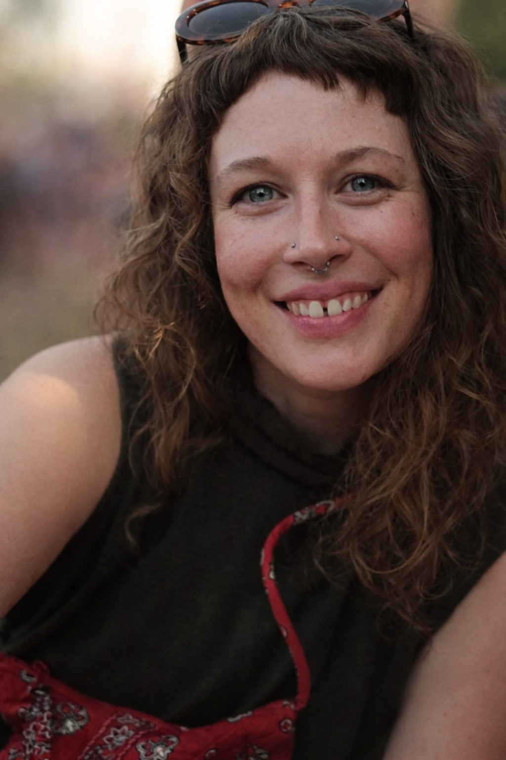 A woman with curly brown hair, blue eyes, and multiple facial piercings, smiling outdoors with a blurred background.