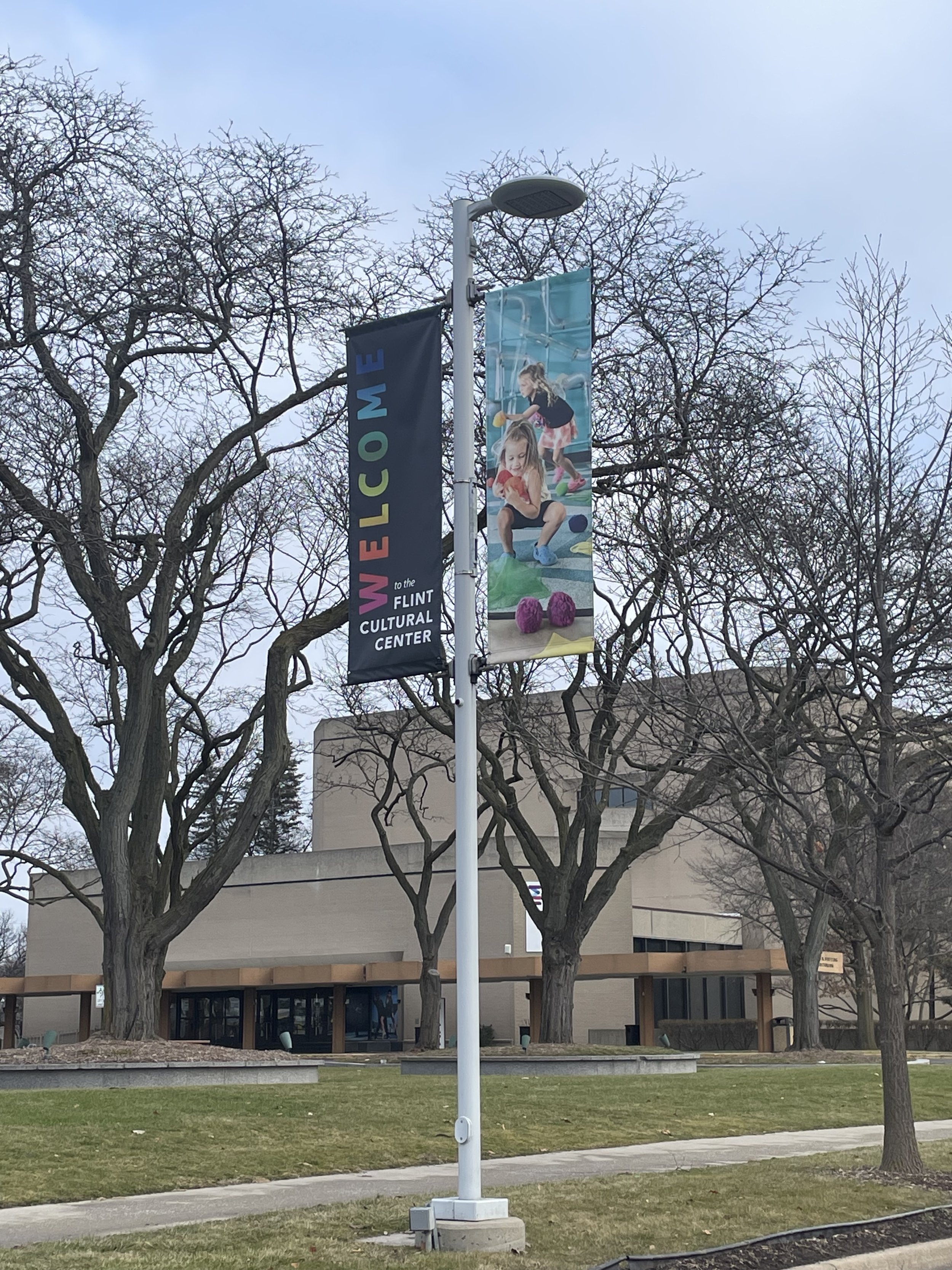 A streetlamp with two banners hanging from it, one reading "WELCOME" and the other featuring an image of two children playing, with trees and a building in the background.