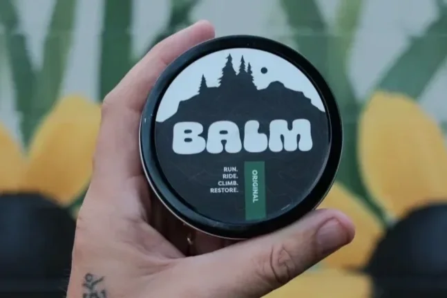 A hand holding a container of BAHM balm, with mountains and trees on the label, and the words "RUN RIDE CLIMB RESTORE" on it.