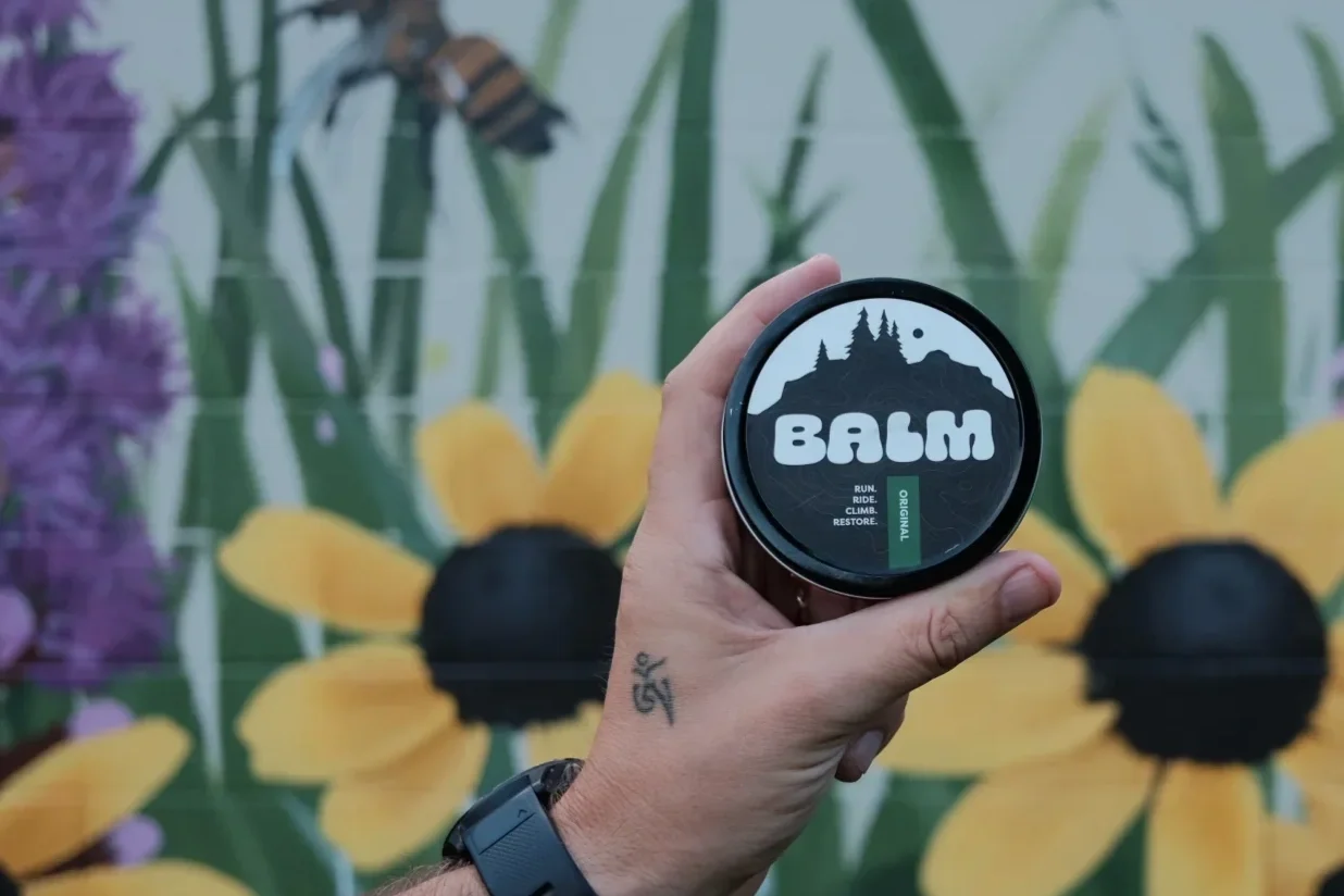 Hand holding a small round container of Balm with a forest mountain logo and the words "Run, Ride, Climb, Restore" against a mural of yellow flowers, green leaves, and a bee.