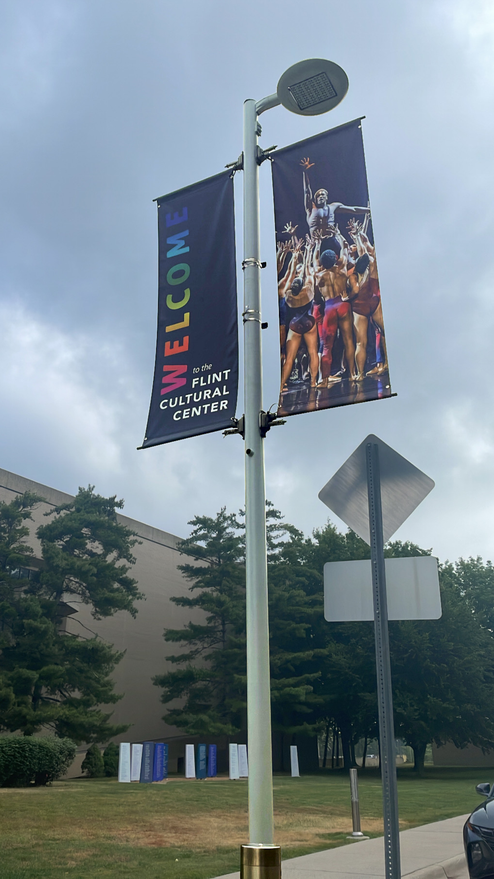 A street lamp with two banners, one says 'Welcome to the Flint Cultural Center' and features a crowd scene with performers, under cloudy skies, with trees and a building in the background.