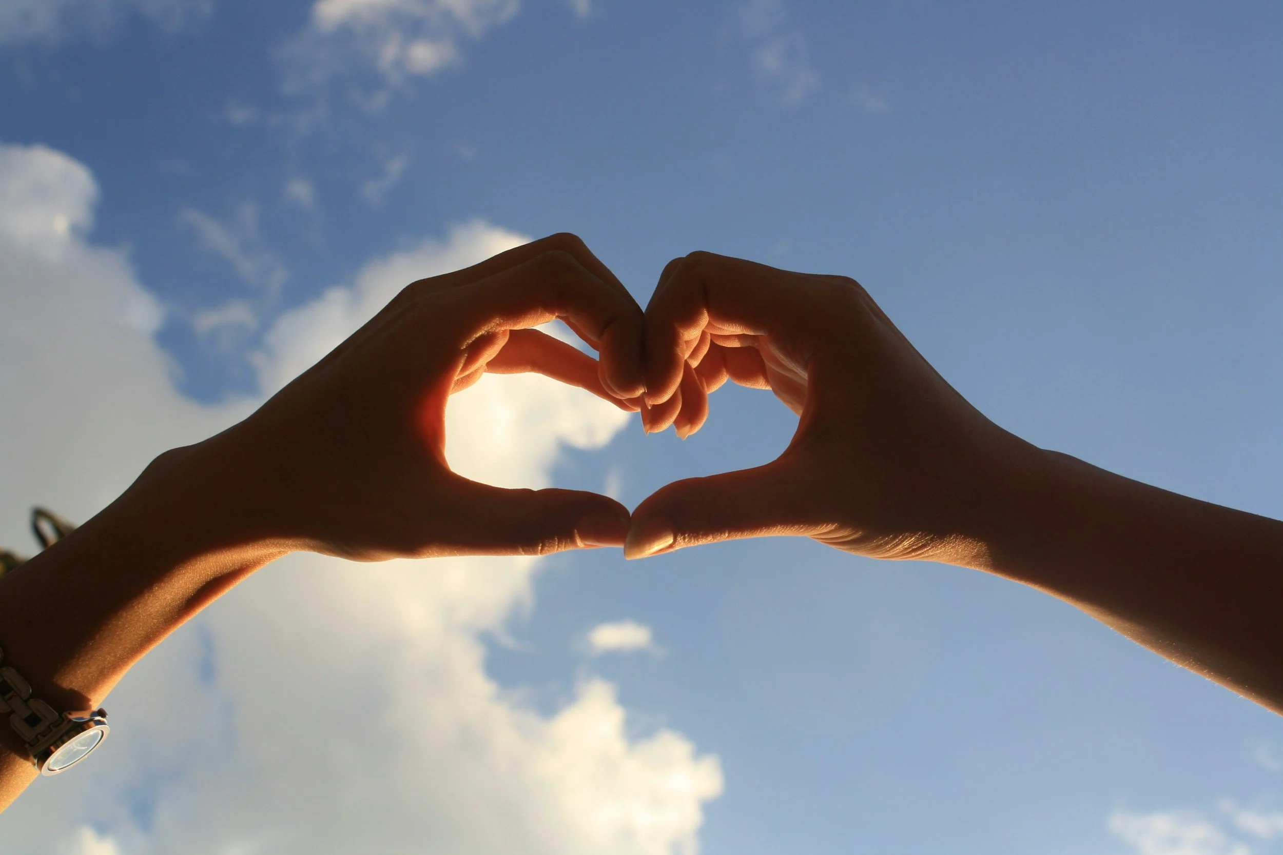 Two hands forming a heart shape against a blue sky with clouds.