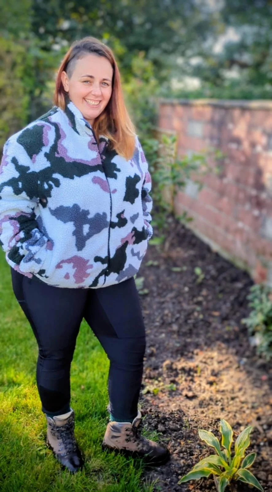A woman standing outdoors in front of a garden with plants and a brick wall, wearing a camouflage jacket and black pants, smiling at the camera.