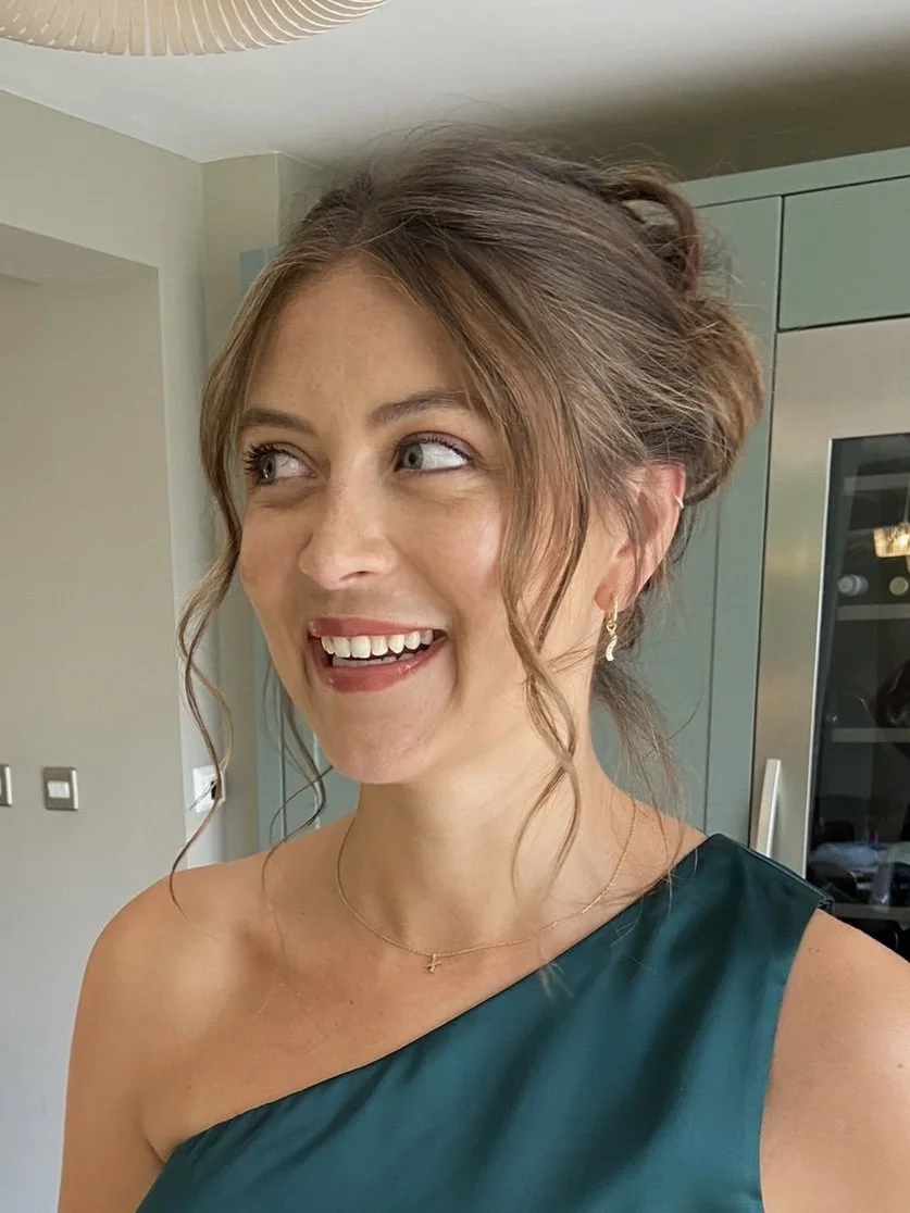 A woman with light skin and brown hair styled in loose curls, smiling and looking to her left. She is wearing a teal one-shoulder dress, gold earrings, and a delicate gold necklace with a small cross pendant. The background includes a kitchen with a 