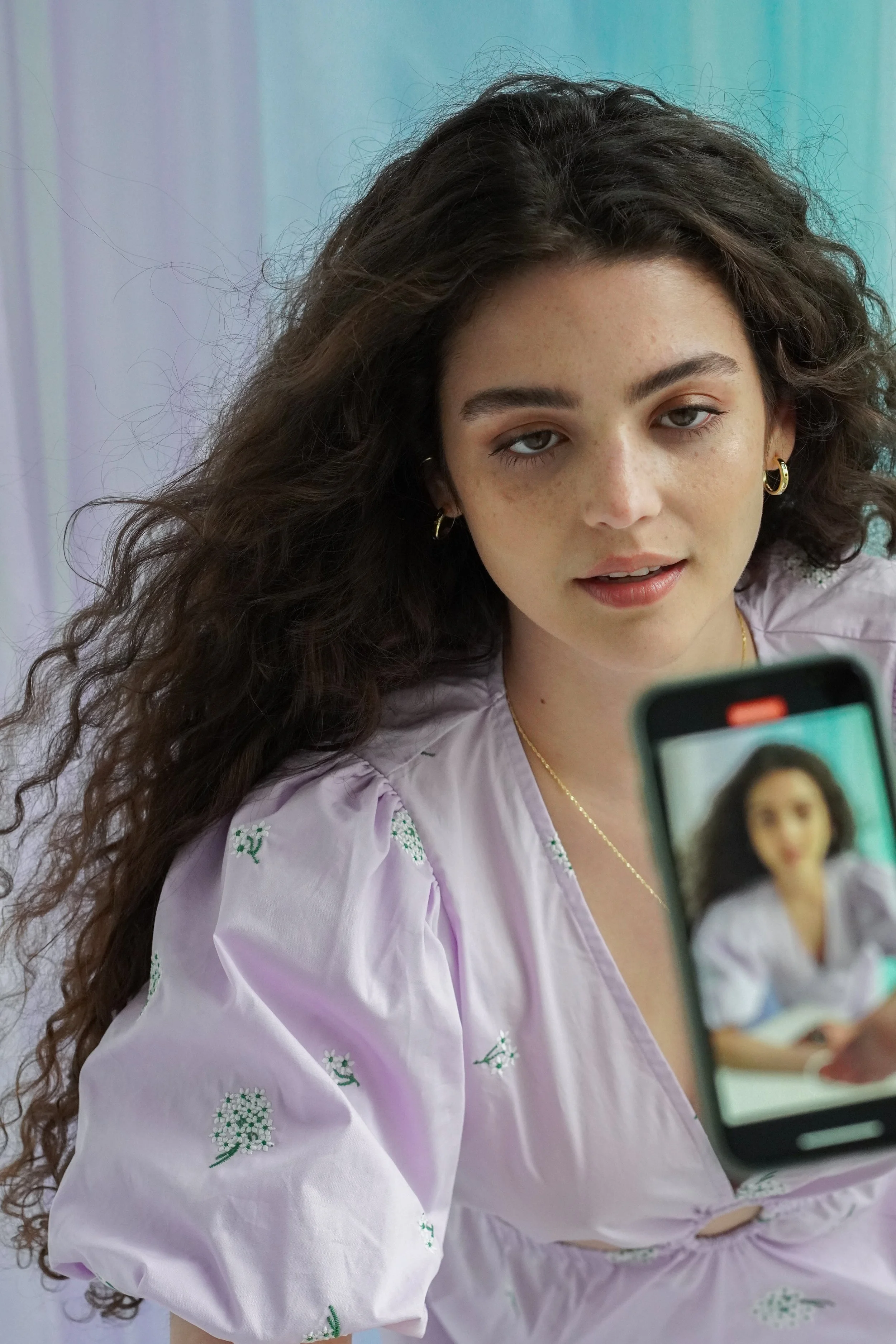 A young woman with long, curly brown hair and light skin taking a selfie with her phone. She is wearing a light purple blouse with embroidered floral patterns and gold hoop earrings. She has freckles and is looking into the camera with a relaxed expr