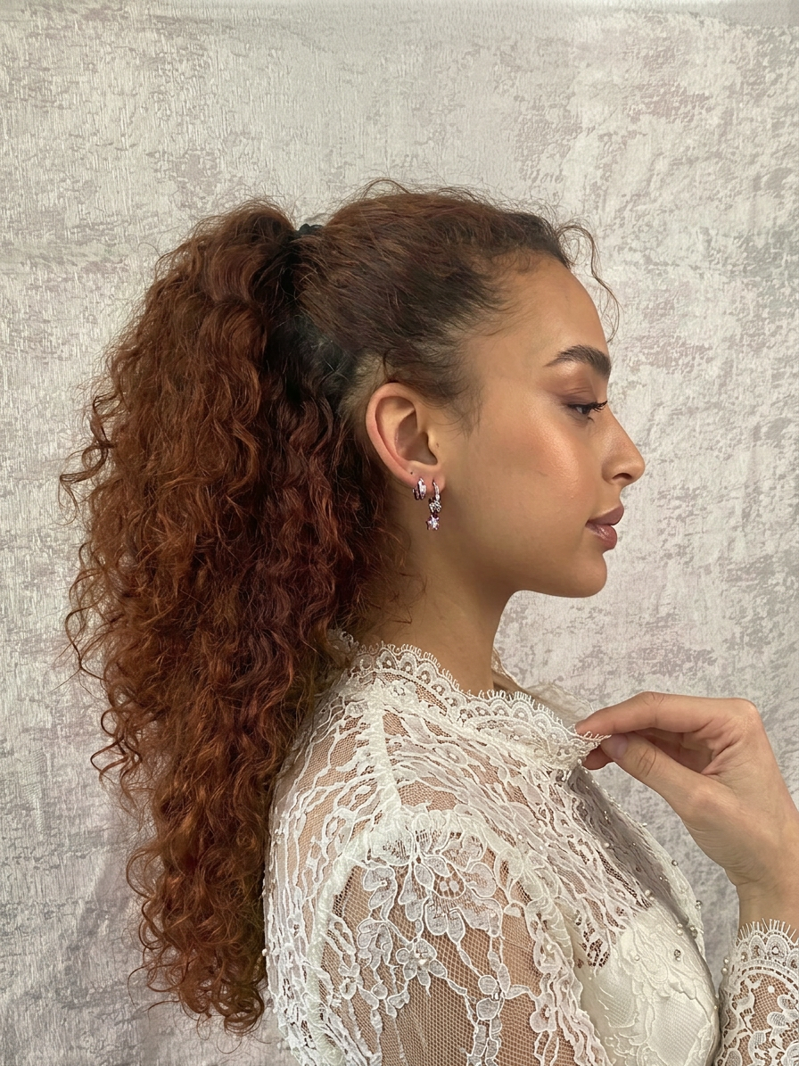 A woman with long, curly red hair styled in a high ponytail, wearing earrings and a white lace top.
