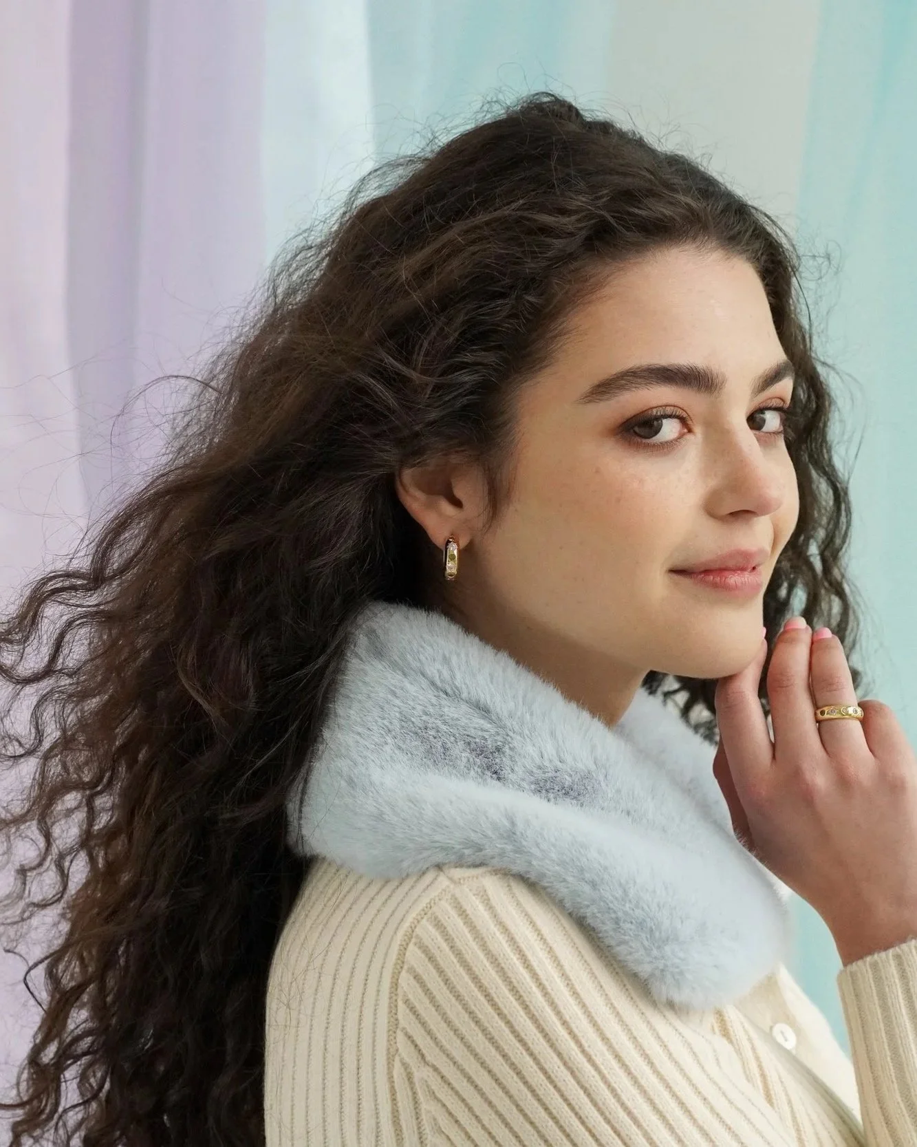 A young woman with curly brown hair wearing a cream-colored sweater with a light blue furry collar, gold hoop earrings, and a gold ring, looking over her shoulder with a soft smile.