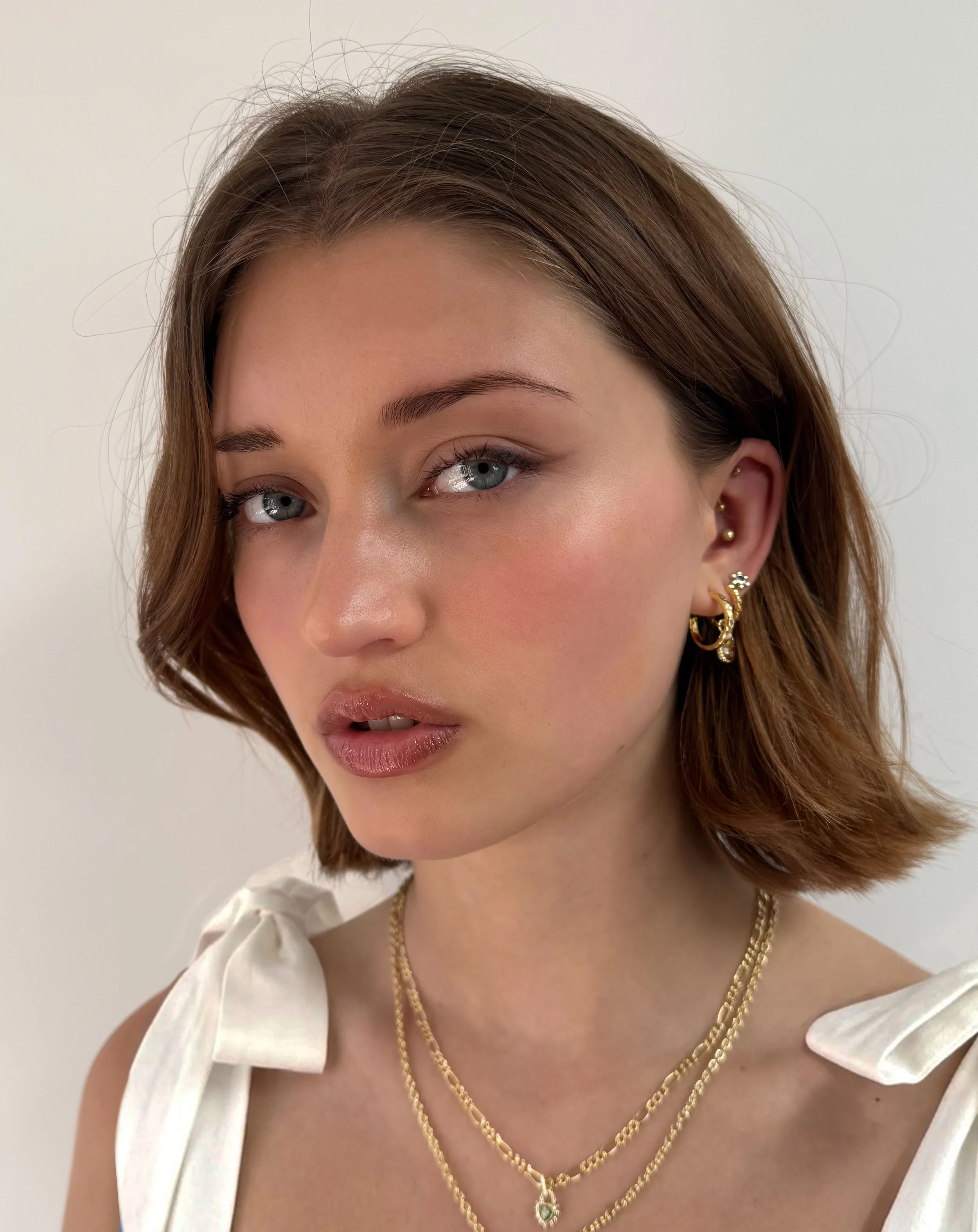 Close-up of a woman with shoulder-length brown hair, blue eyes, and light skin, wearing layered gold necklaces, multiple earrings, and a white top with bows on the shoulders.