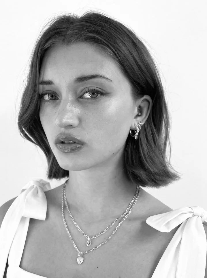 Black and white portrait of a young woman with short, wavy hair. She has multiple earrings, layered necklaces, and is wearing a top with bow-tied shoulder straps.