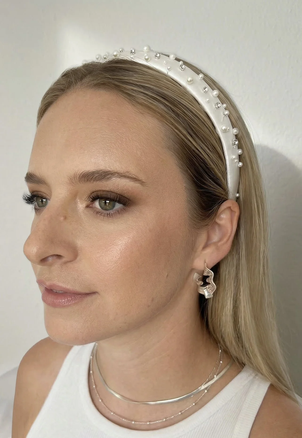 A woman with blonde hair wearing a white pearl-embellished headband, layered silver necklaces, and geometric earrings, facing slightly to the right.