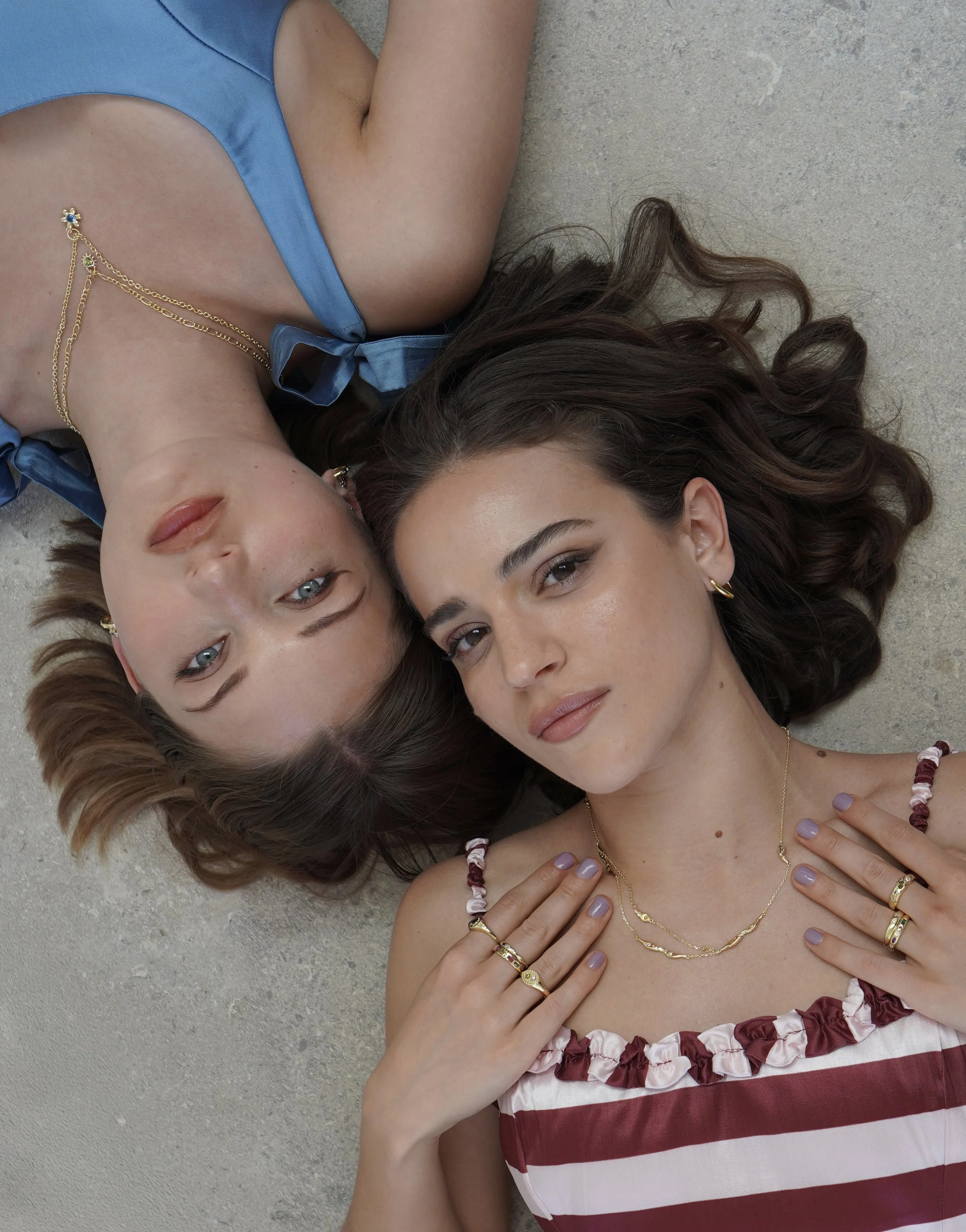 Two women with colorful jewelry lie on a concrete surface, facing upward. One woman has dark brunette hair and the other has light brown hair with highlights. Both are wearing casual tops and jewelry, and look directly at the camera.