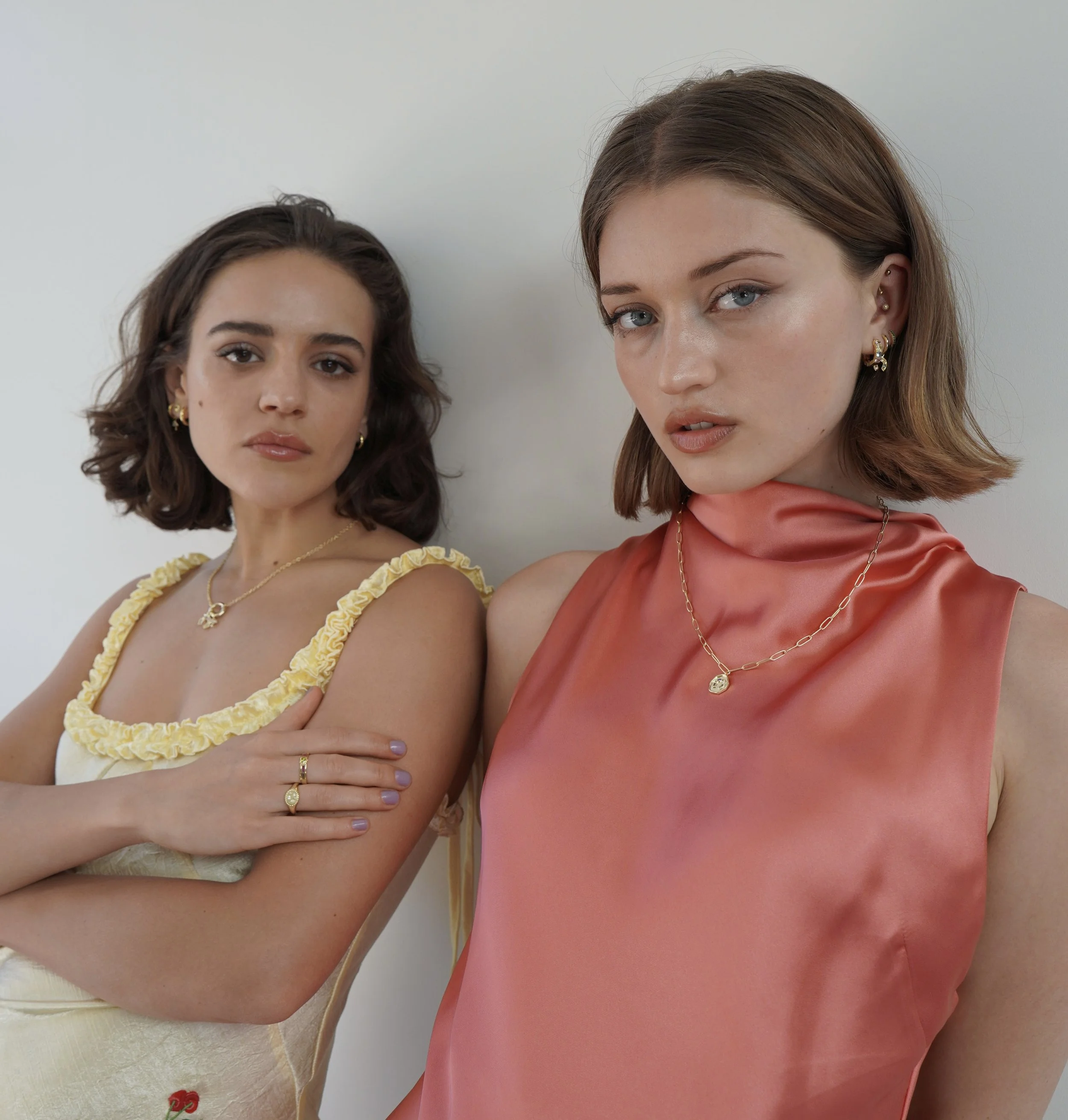 Two women with shoulder-length brown hair, wearing jewelry, standing close together against a plain white wall.
