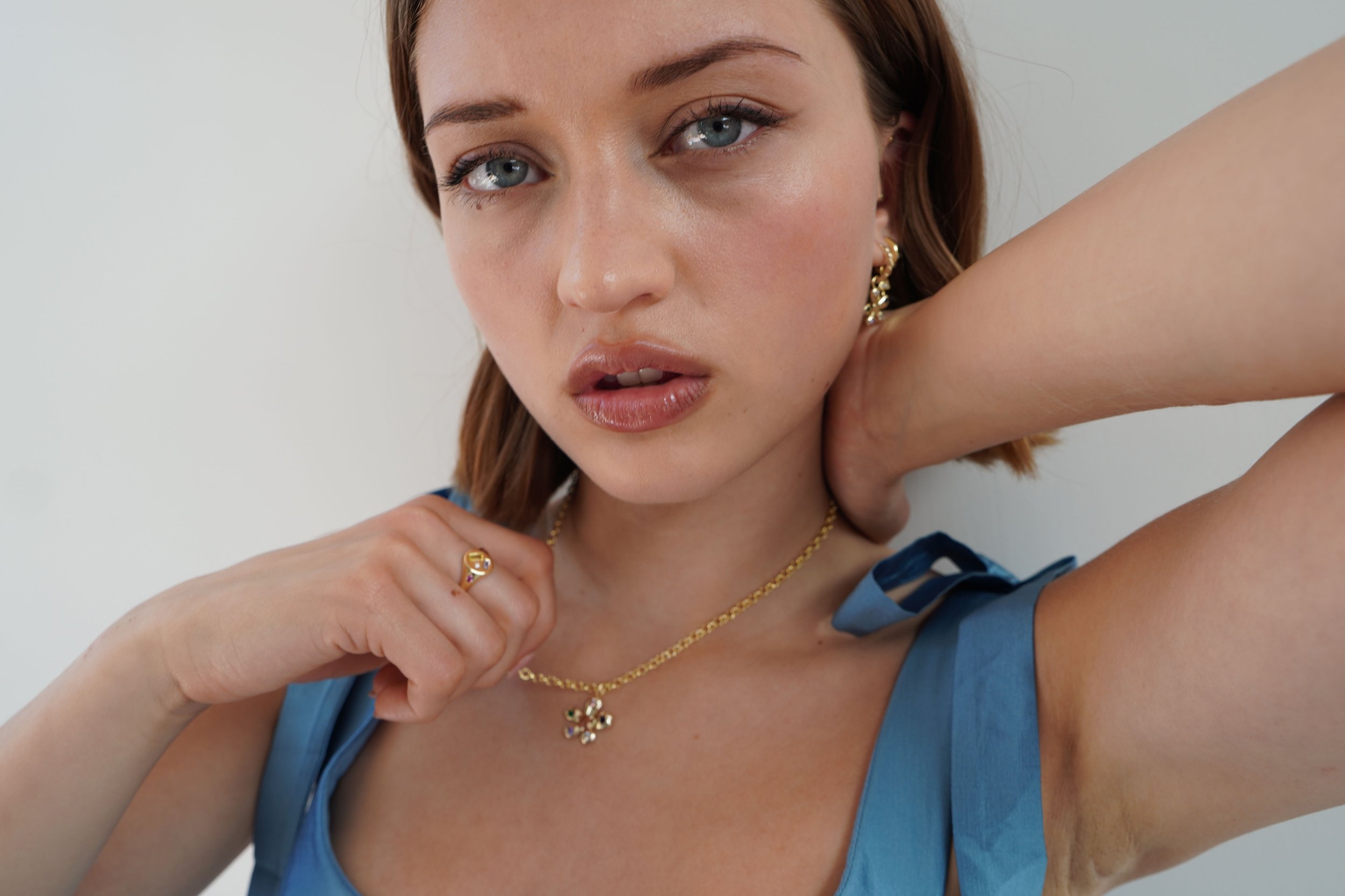 A woman with blue eyes and fair skin posing with her hand near her neck against a plain background. She is wearing gold jewelry including earrings, a necklace with a butterfly pendant, and a ring. She has makeup with natural tones and wears a blue sl