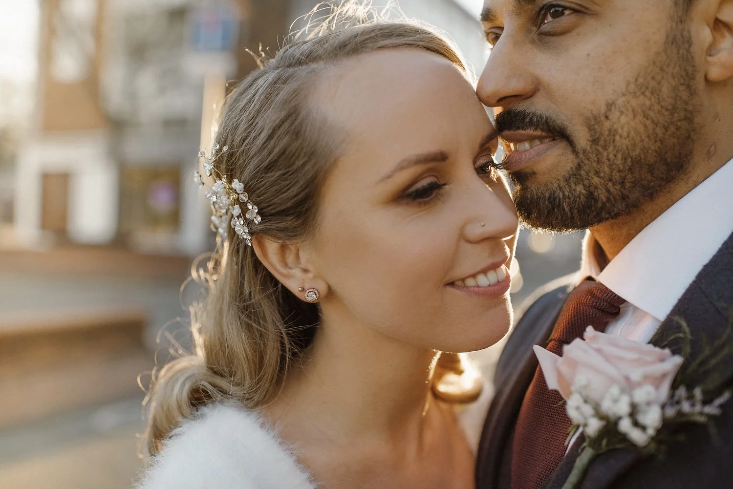 Close-up of a smiling bride and groom in a romantic outdoor setting, with the bride wearing floral hair accessories and earrings, and the groom in a dark suit with a boutonniere.