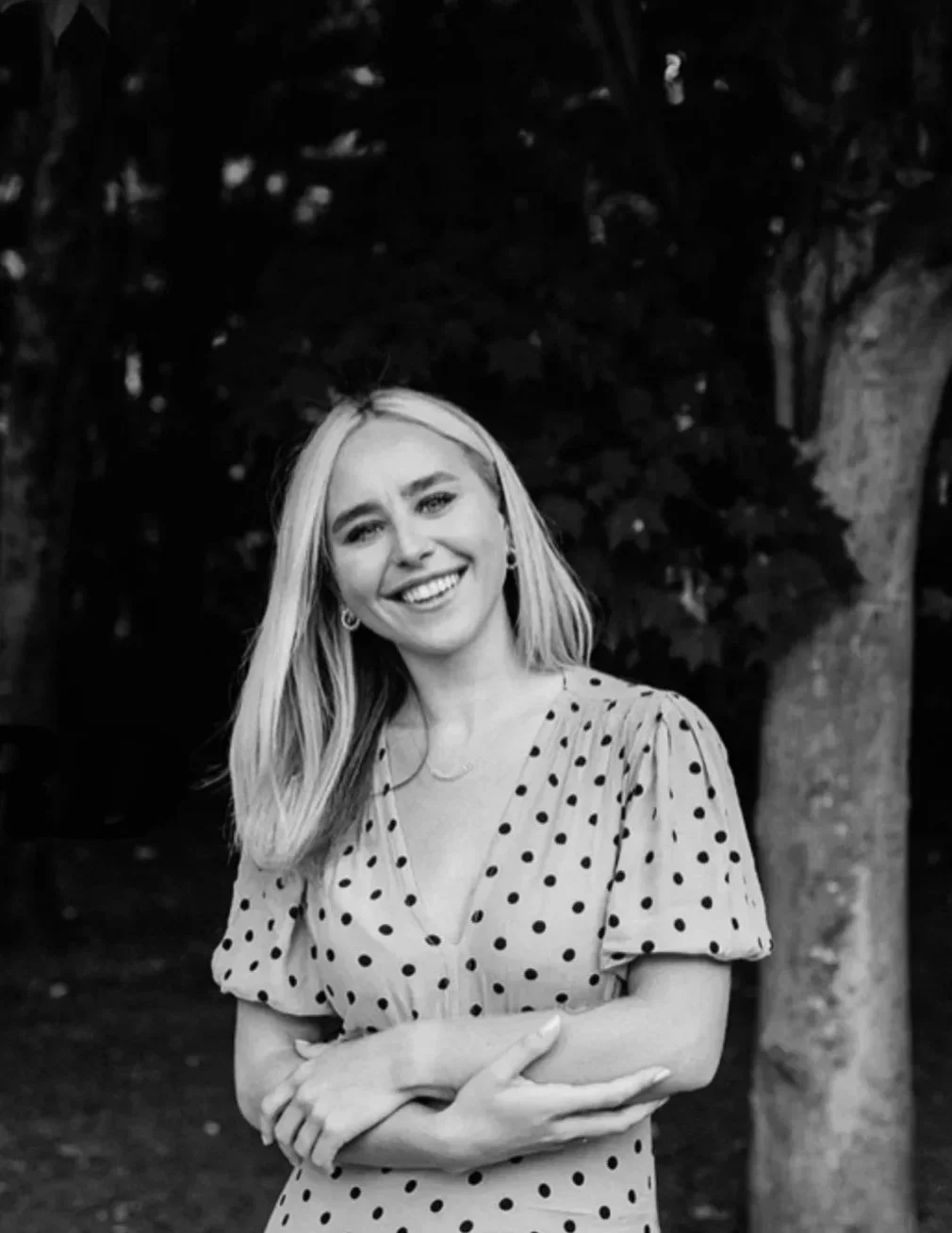 A young woman with blonde hair smiling and standing outdoors in front of trees, wearing a polka dot dress.