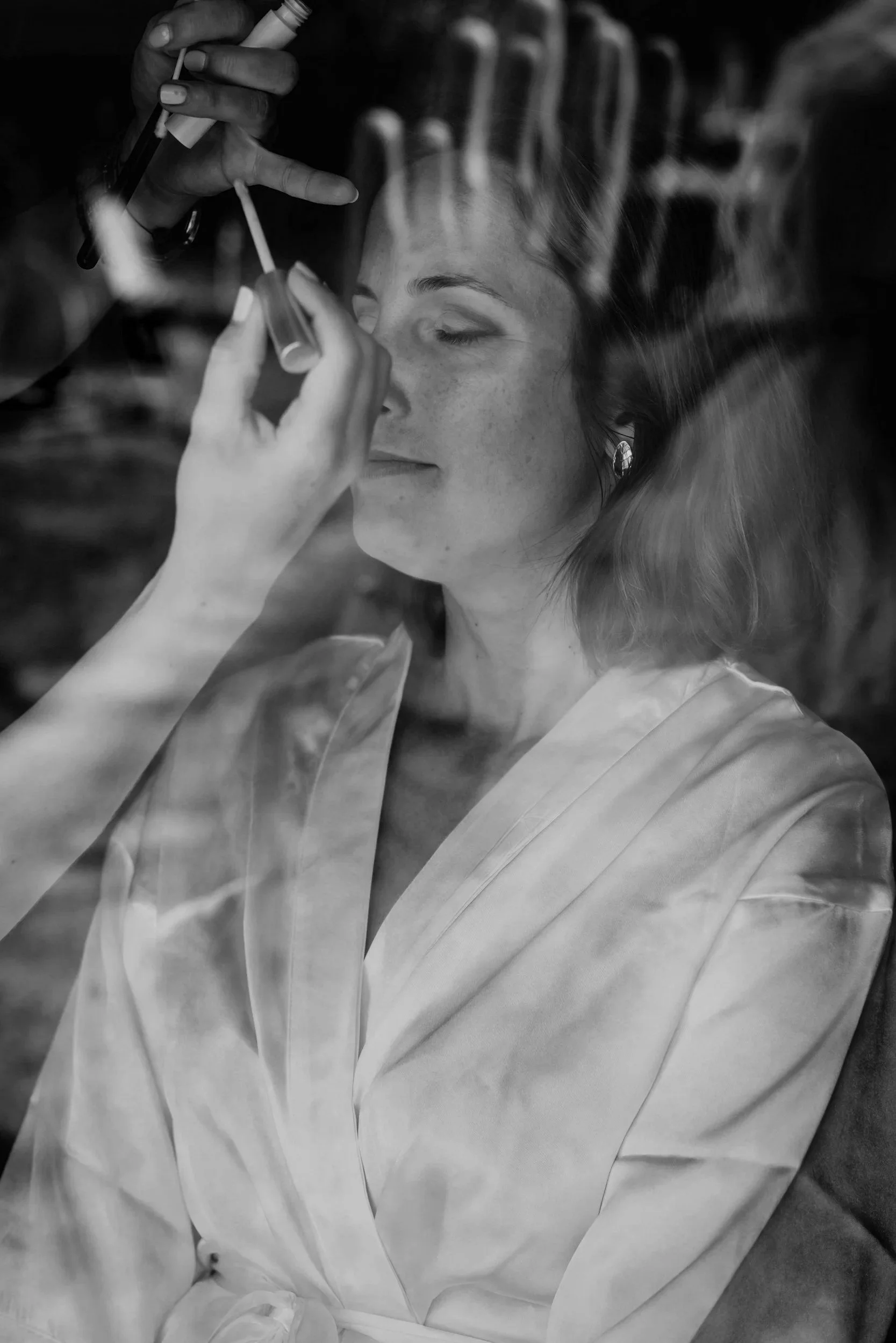 A woman with closed eyes, wearing earrings and a light-colored robe, is getting makeup applied by a person with hands holding a makeup brush and a small container, seen through a reflective surface.