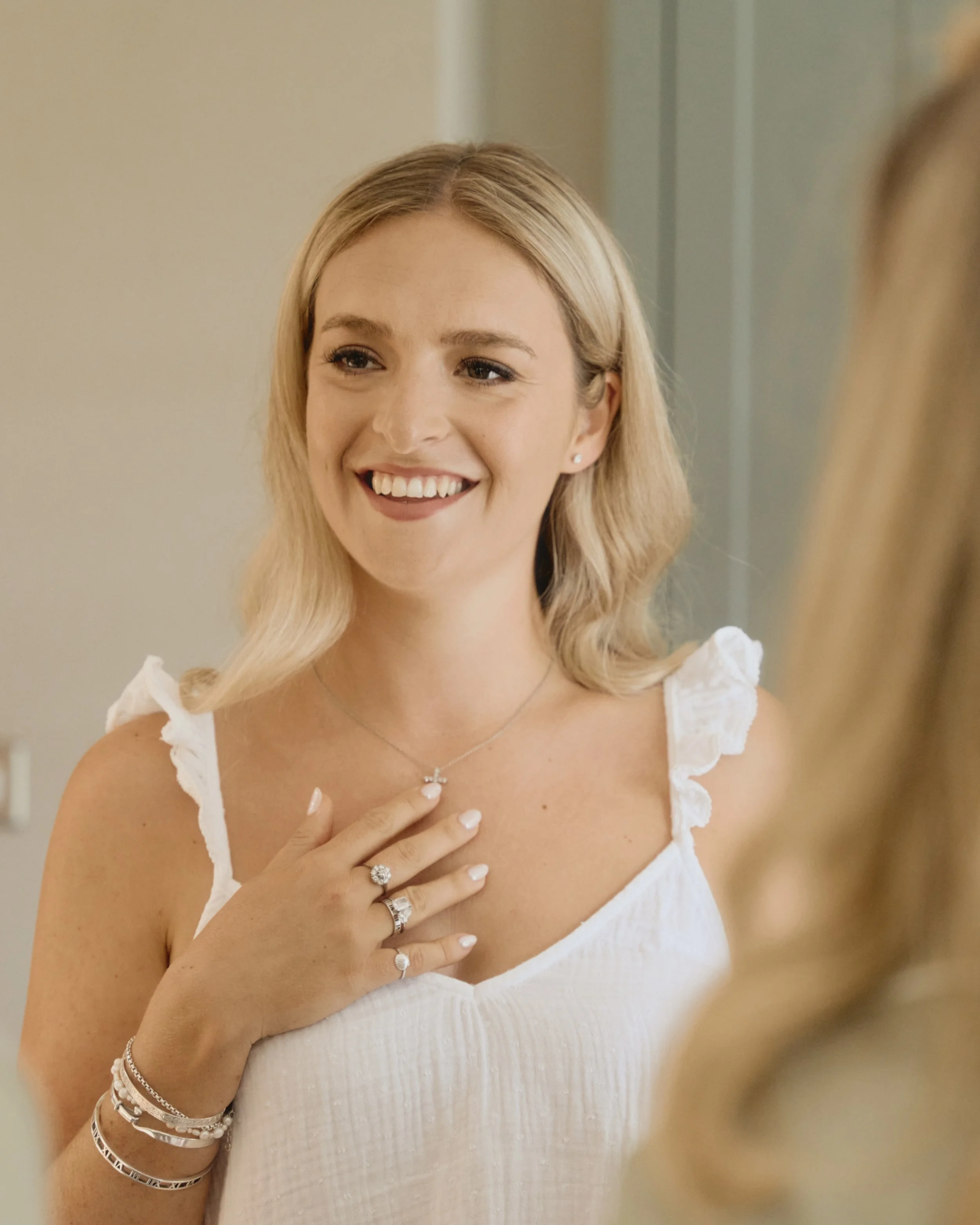 A young woman with blonde hair smiling and showing her jewelry, including rings, bracelets, a necklace, and earrings, while touching her chest.