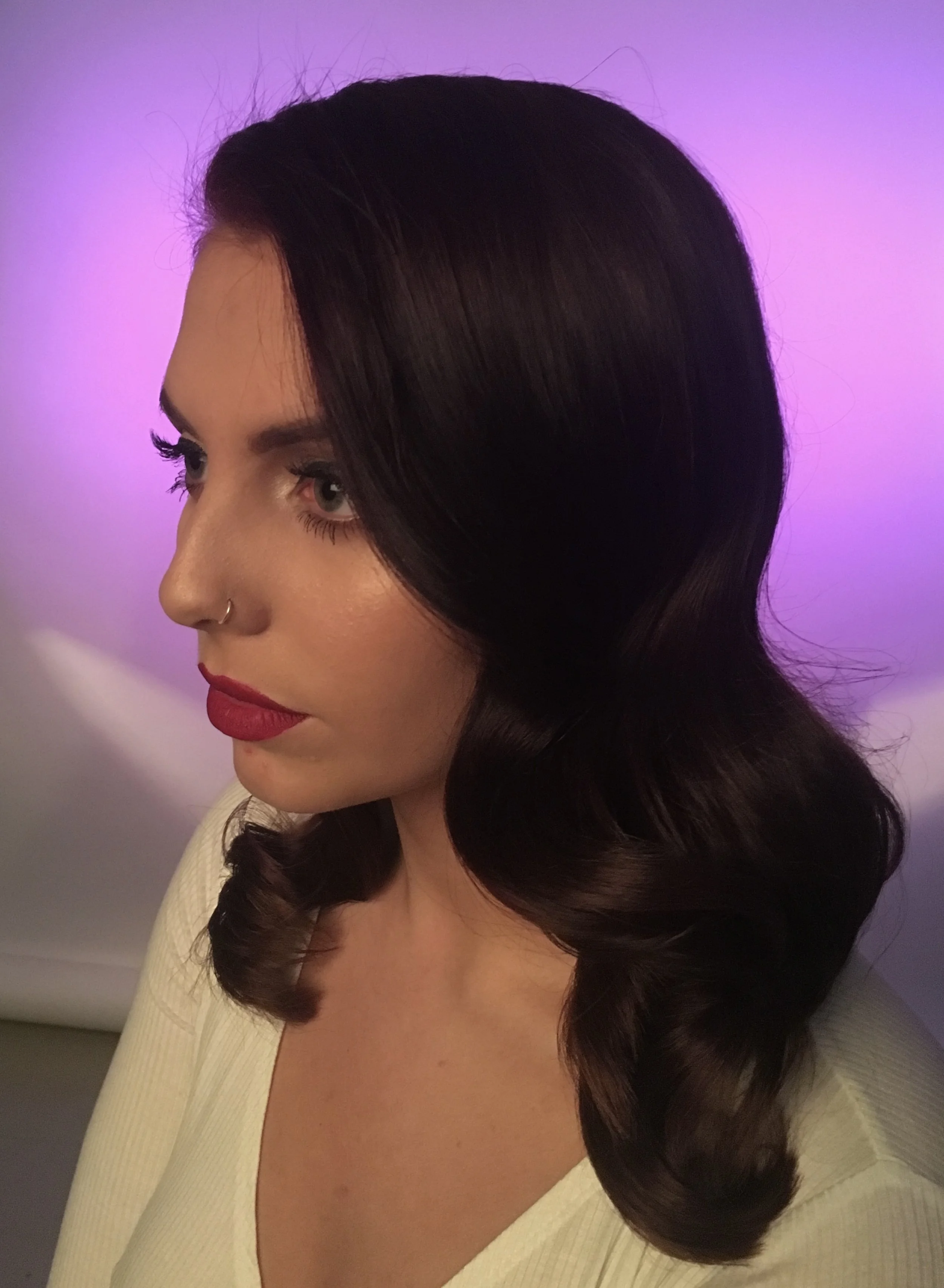 A woman with dark brown, wavy hair, wearing red lipstick and eyeliner, is facing to the left in front of a purple and pink gradient background.
