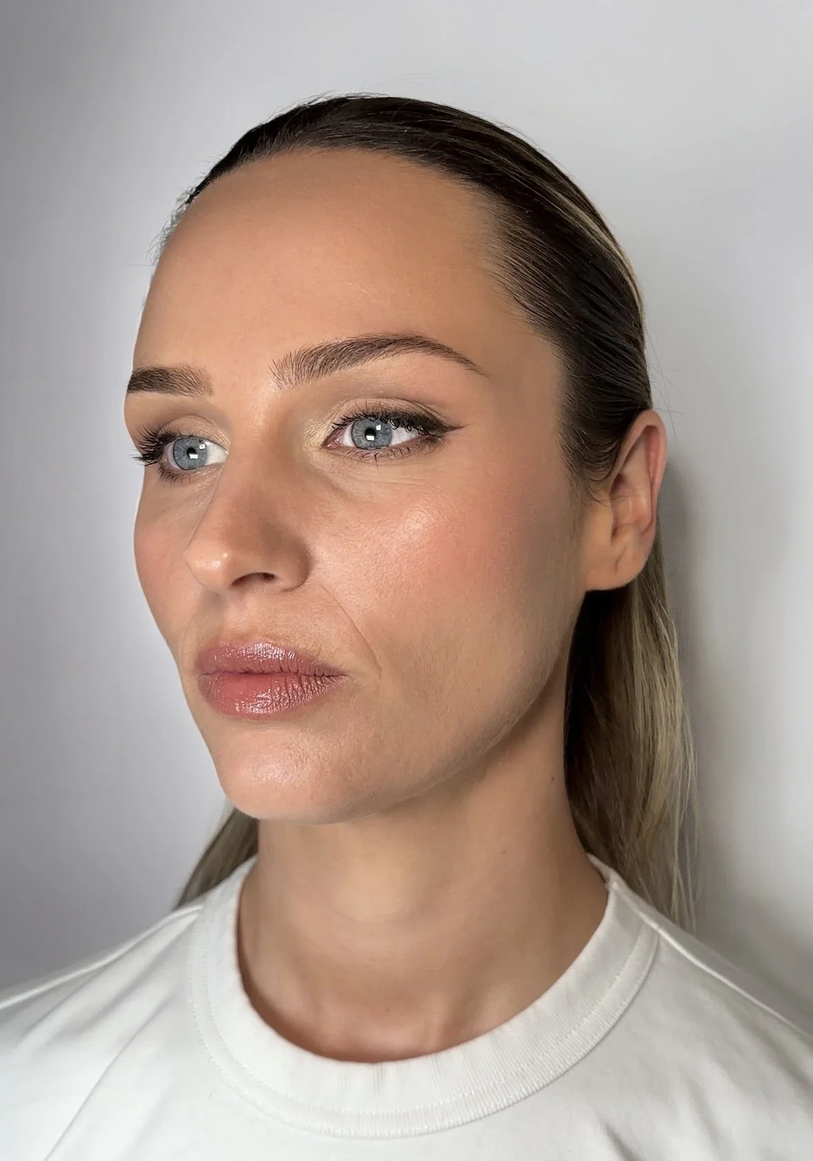 A close-up portrait of a woman with fair skin, blonde hair, and blue eyes, wearing natural makeup and a light-colored shirt, against a plain white background.
