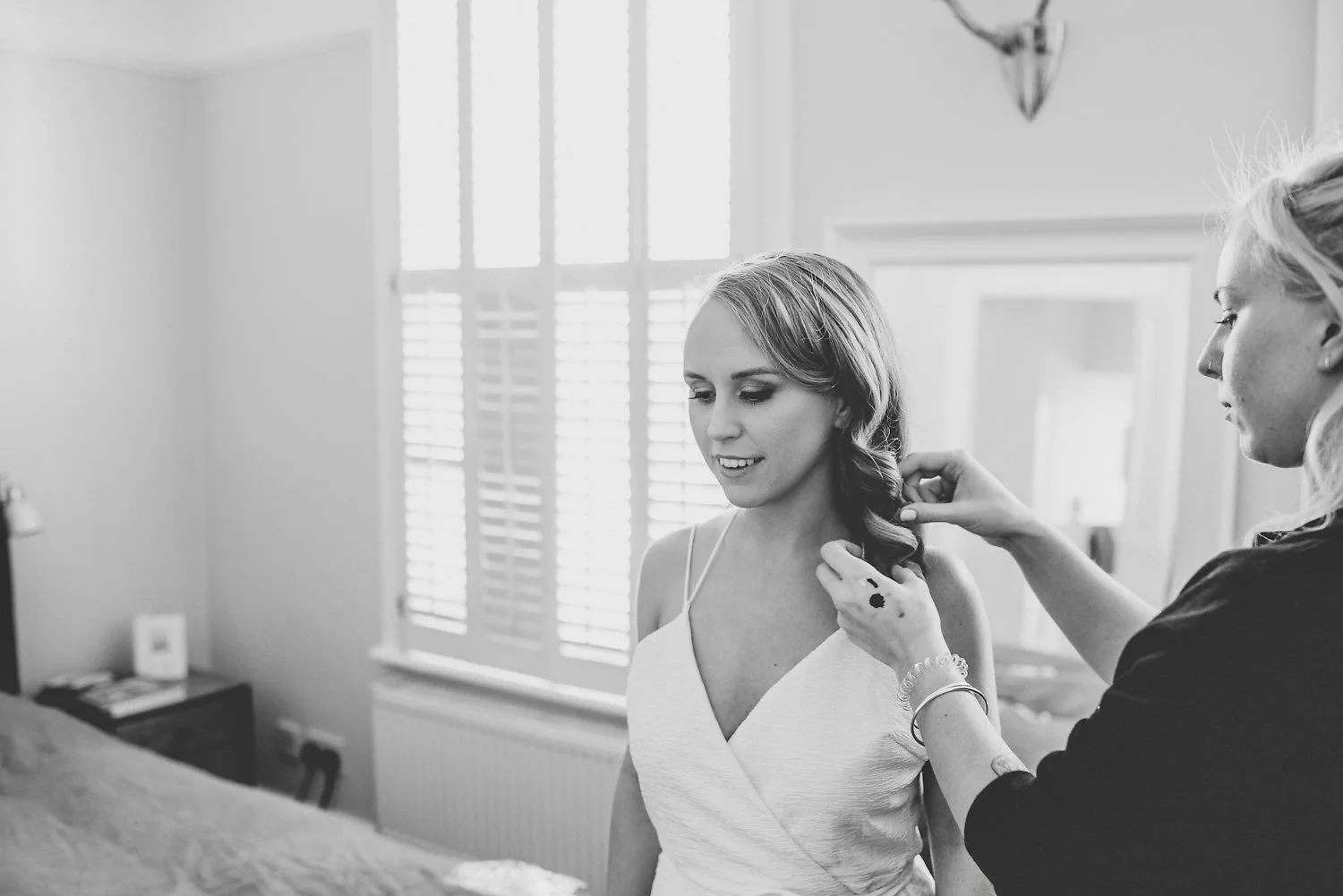 A woman in a sleeveless dress is having her hair styled by another woman in a black top. They are in a bright room with a window covered by shutters. The woman getting her hair styled is smiling slightly.