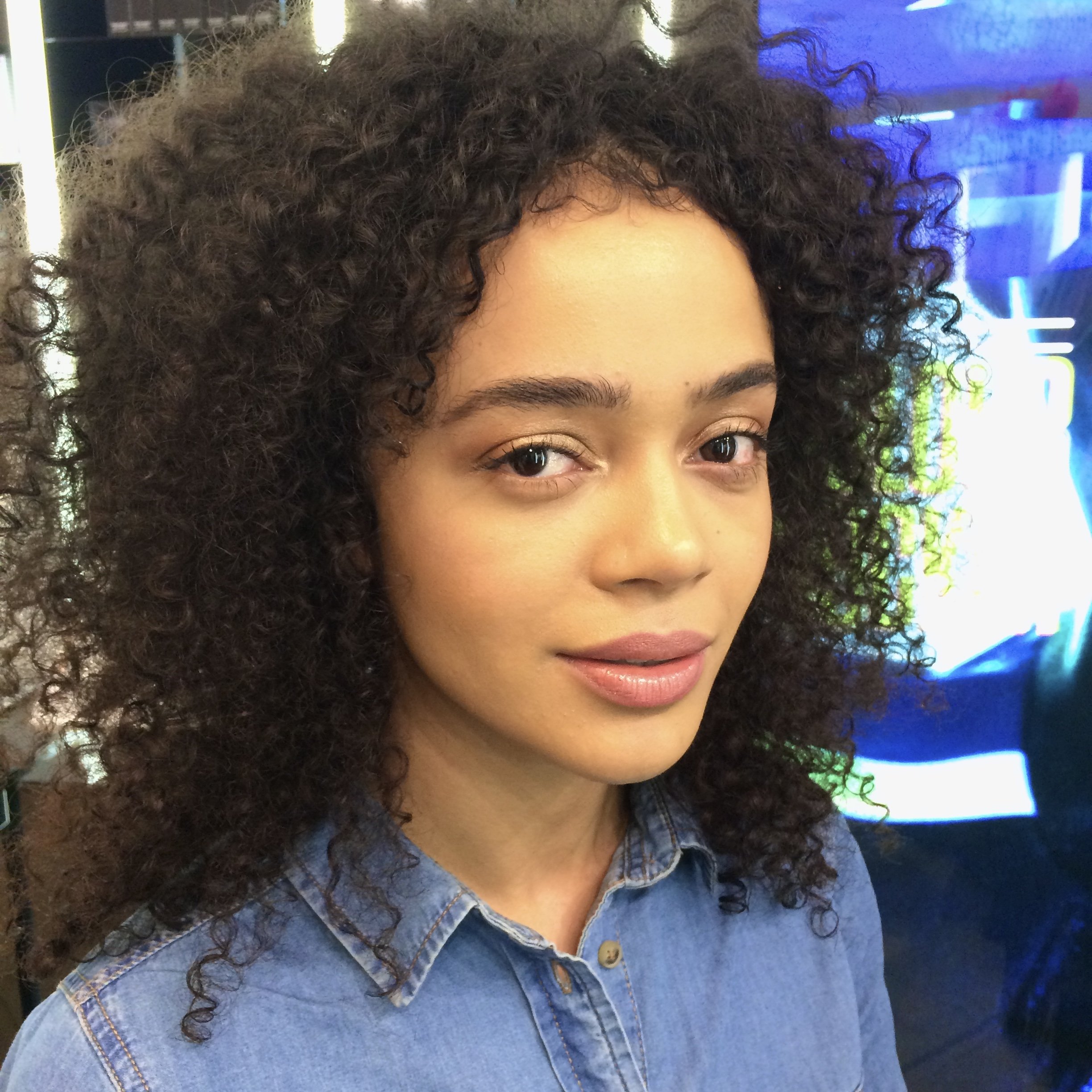 Close-up of a young woman with curly dark hair, wearing a denim shirt, in an indoor setting with a colorful, illuminated background.