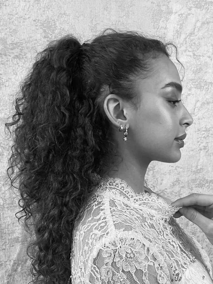 A black-and-white portrait of a woman with curly hair tied in a high ponytail, wearing lace earrings and a lace top, standing against a textured wall, in profile view.