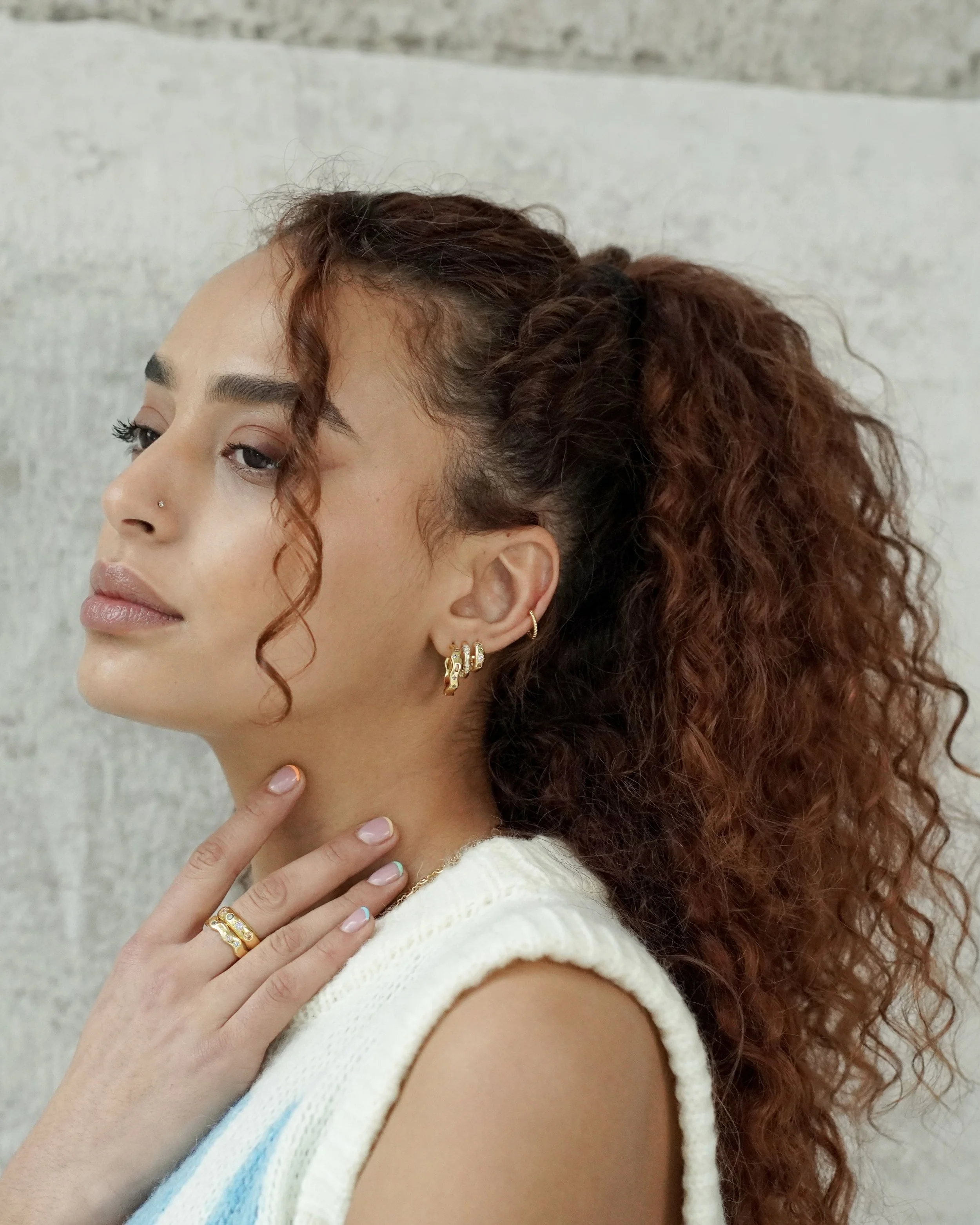 A woman with curly red hair and gold jewelry, including earrings and rings, touches her neck with her left hand, posed against a textured gray background.