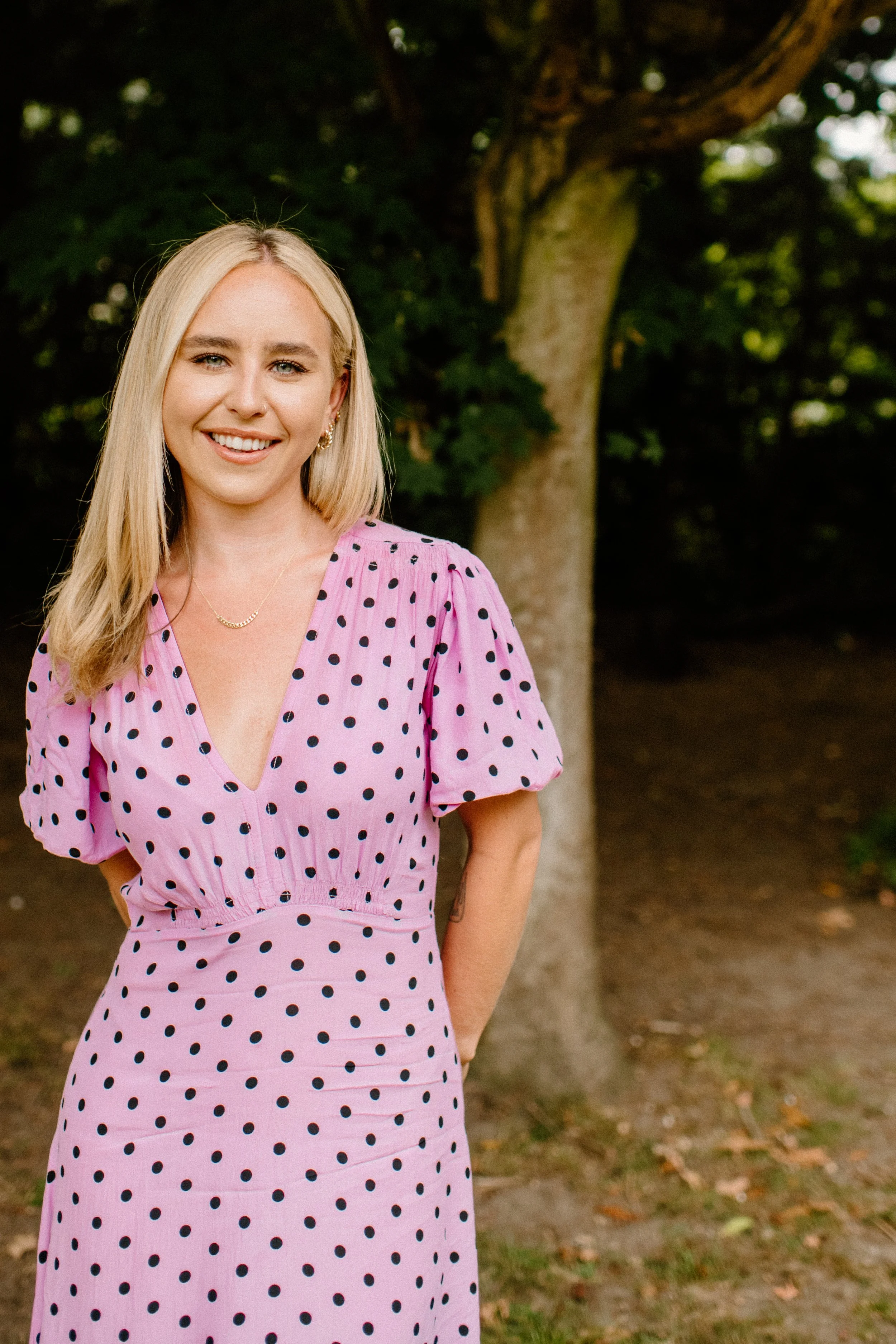 A young woman with blonde hair in a pink polka dot dress standing outdoors near a tree, smiling at the camera.