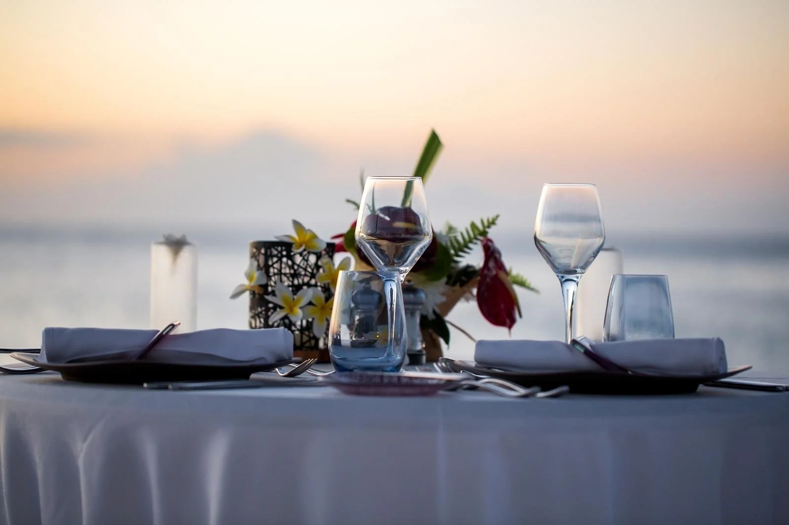 Elegant dining table set outdoors with wine glasses, plates, and silverware, overlooking a body of water at sunset with a floral centerpiece.