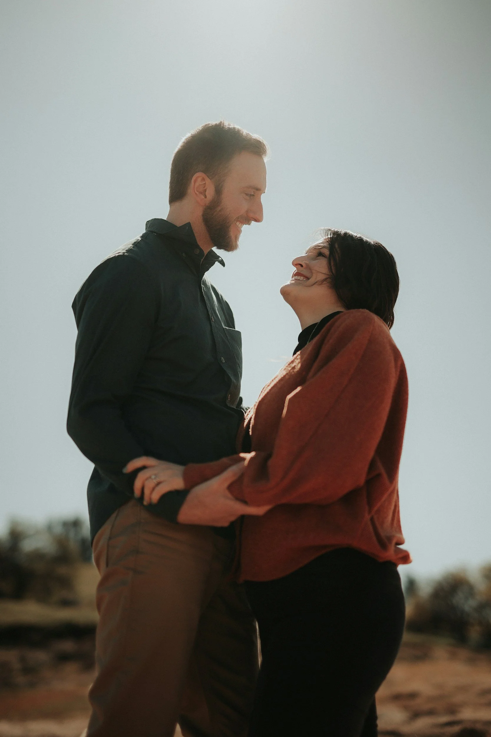 A man and a woman standing close together outdoors, smiling and looking into each other's eyes.