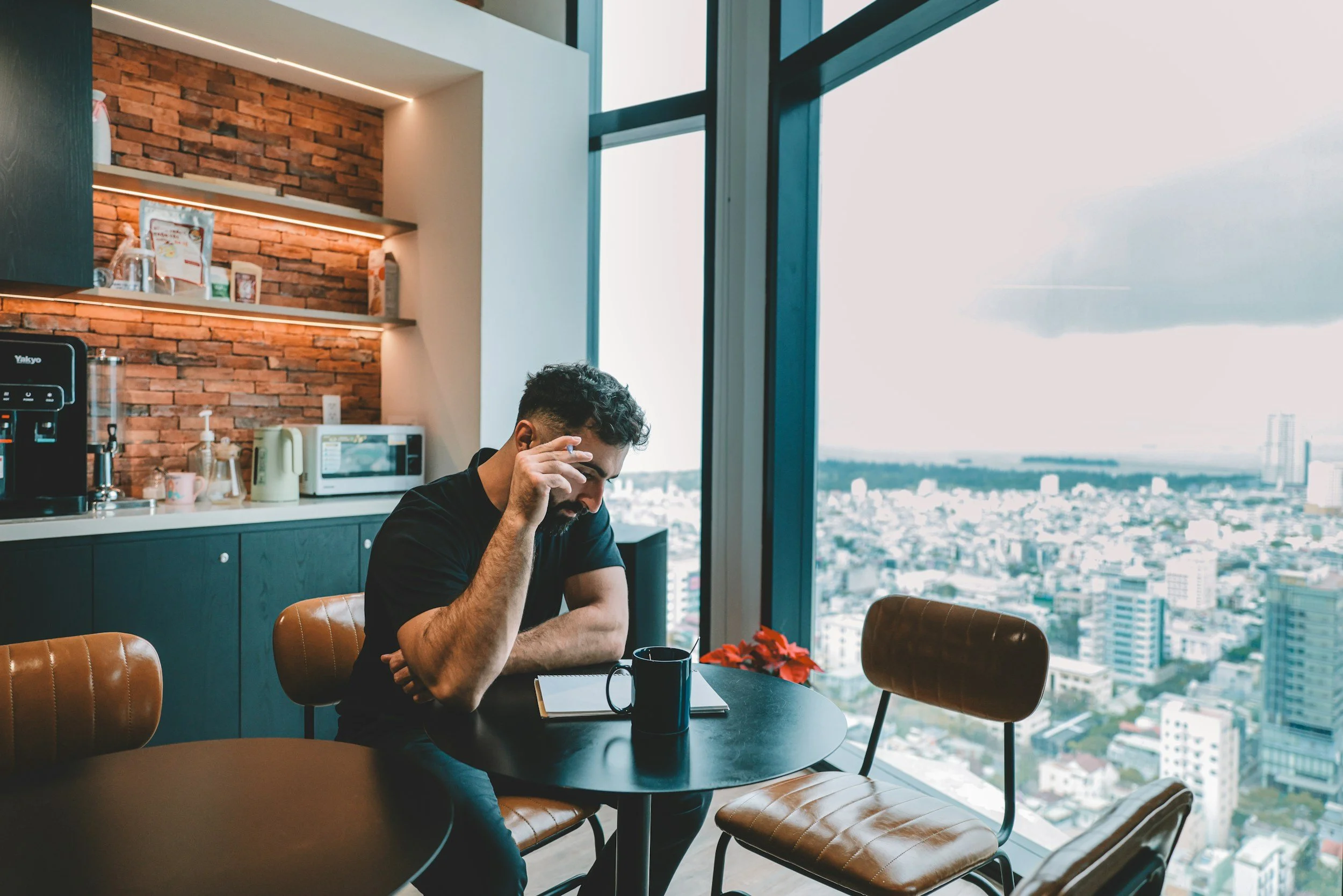 A man sitting at a black round table in a high-rise apartment or office with a city view through large windows. The table has a notebook, a black mug, and a red poinsettia plant. The background includes a kitchen with brick wall, shelves, a microwave, and various appliances.