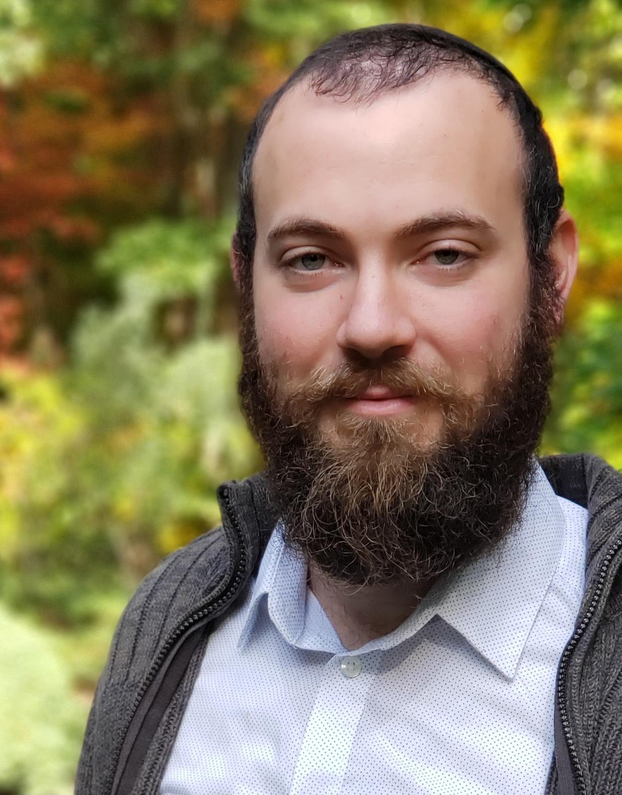 A man with a full beard and short hair smiling outdoors with autumn foliage in the background.