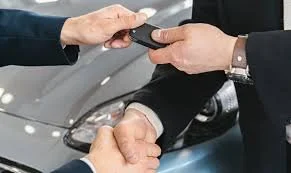 Two people shaking hands with a car in the background, one person hand giving a key to the other.