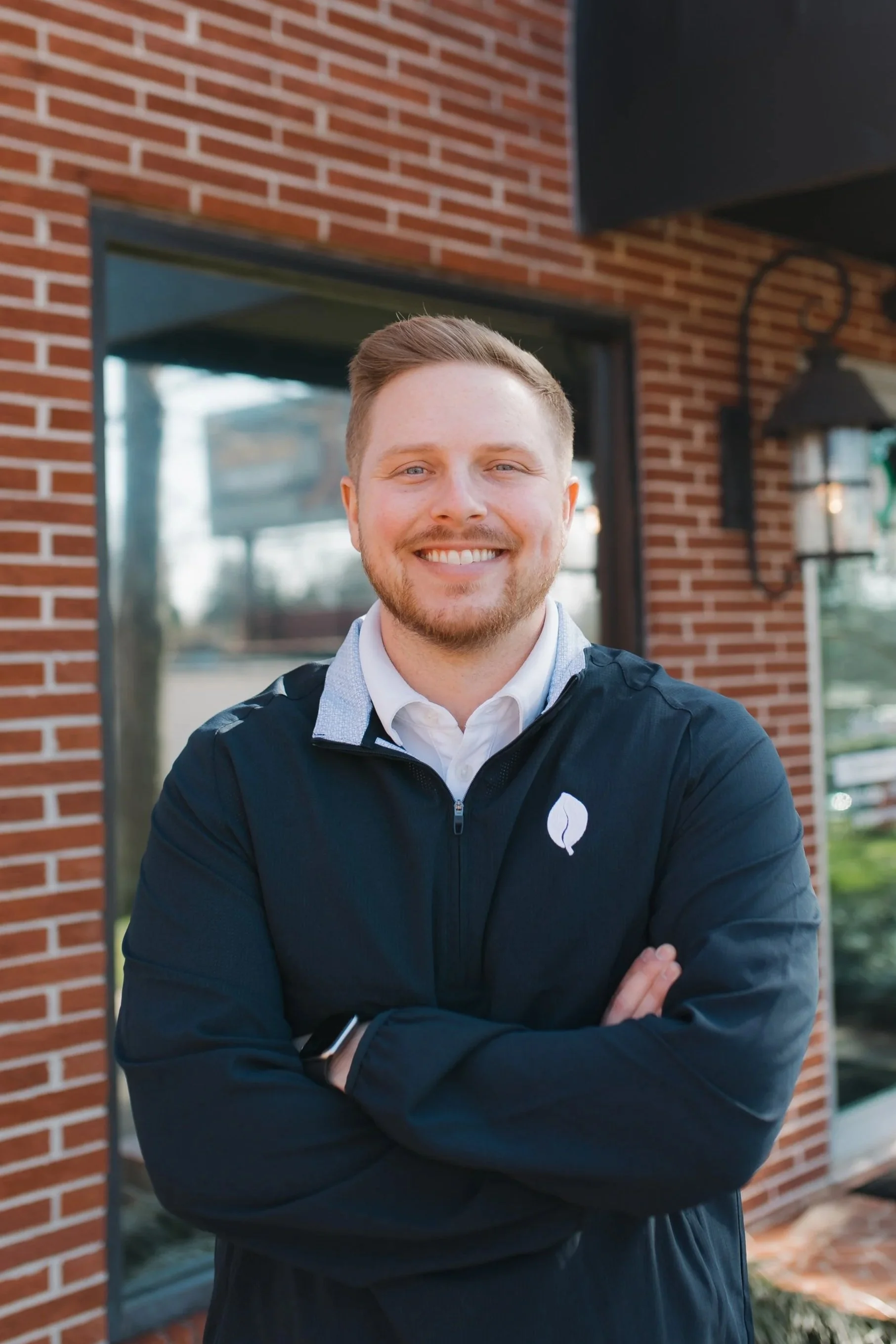 A smiling young man with short reddish hair and a beard standing outside in front of a brick building, wearing a black jacket with a white logo on the chest and a white collared shirt underneath.