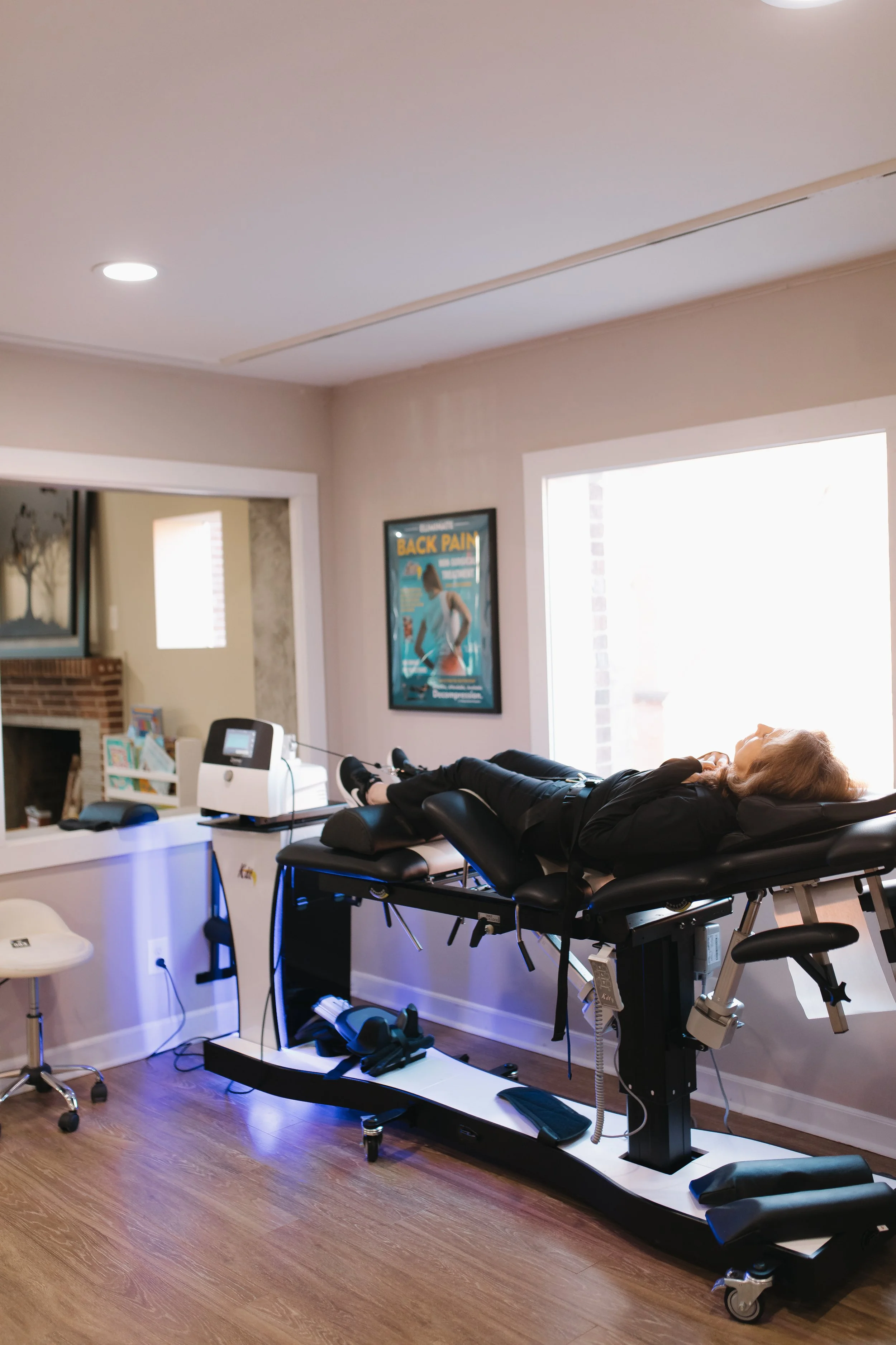A woman lying on a spinal decompression therapy table in a chiropractic office with a large window, a framed poster on the wall about back pain, and equipment nearby.