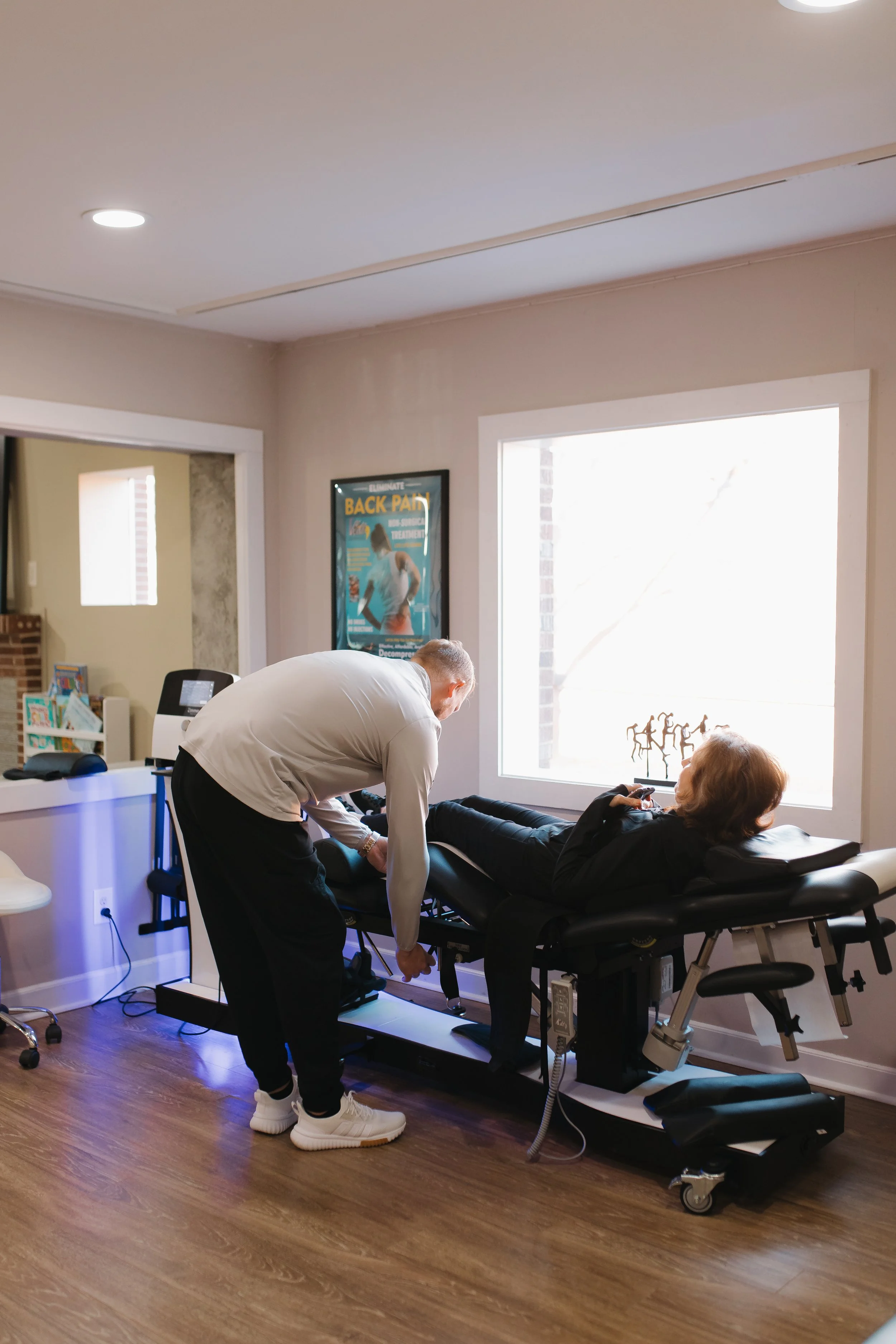 Medical professional assisting a patient lying on a spinal decompression therapy table in a chiropractic office with a large window, framed artwork, and medical equipment.