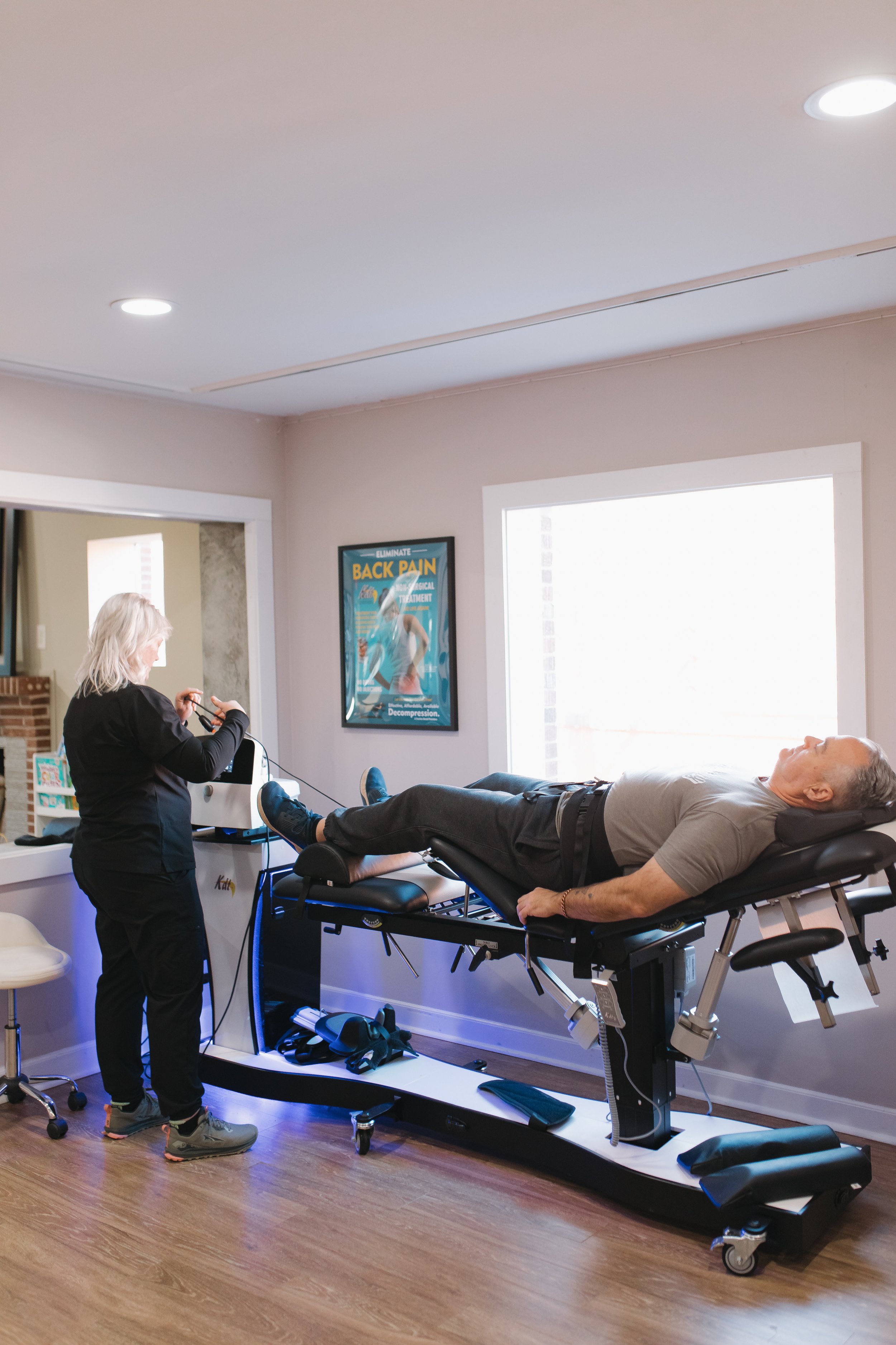 A patient lying on a spinal decompression therapy  table, connected to a machine, in a clinic room with a window and a poster on back pain on the wall.