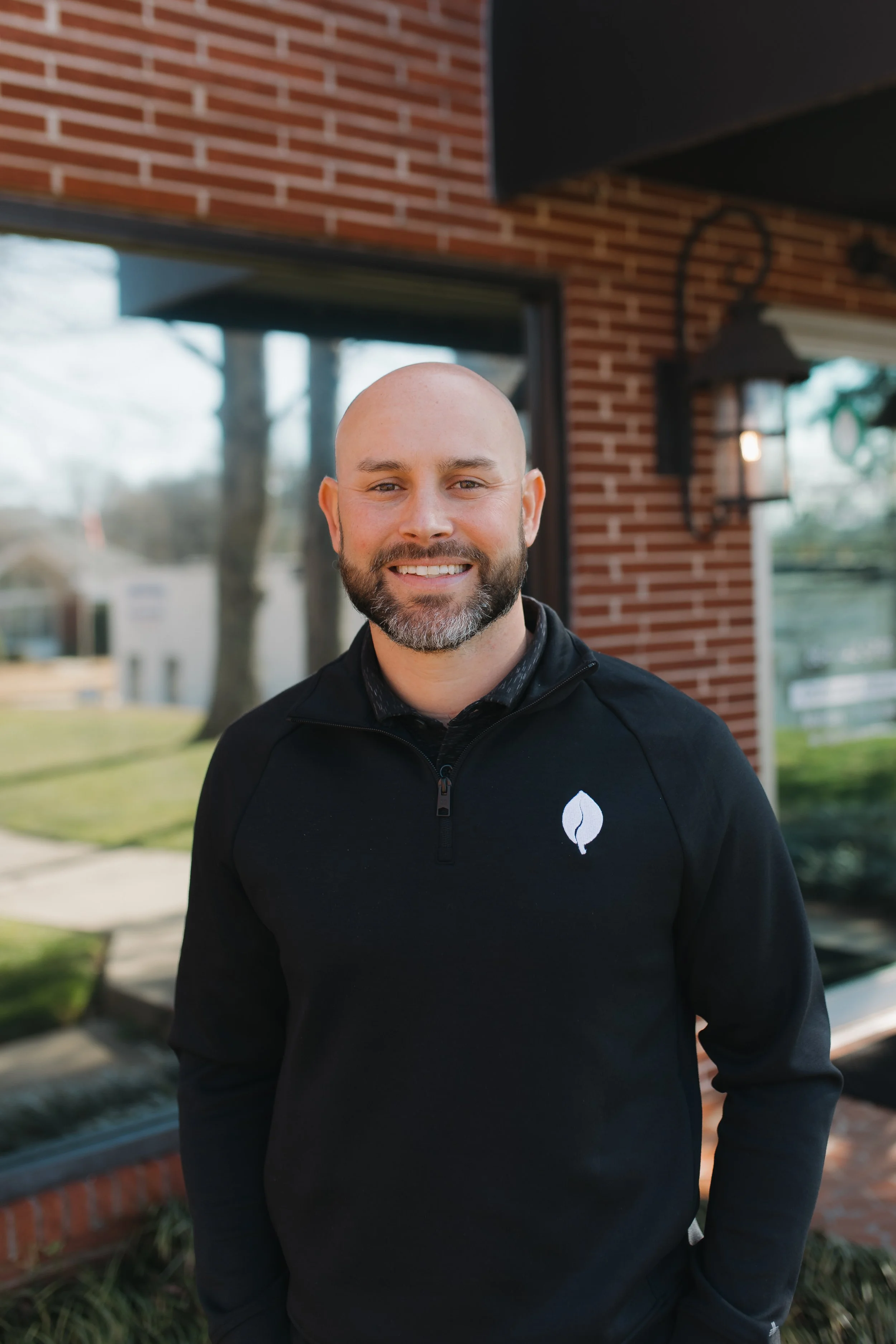 A smiling Chiropractor with a beard and shaved head stands outside near a brick building, wearing a black jacket with a white leaf logo.
