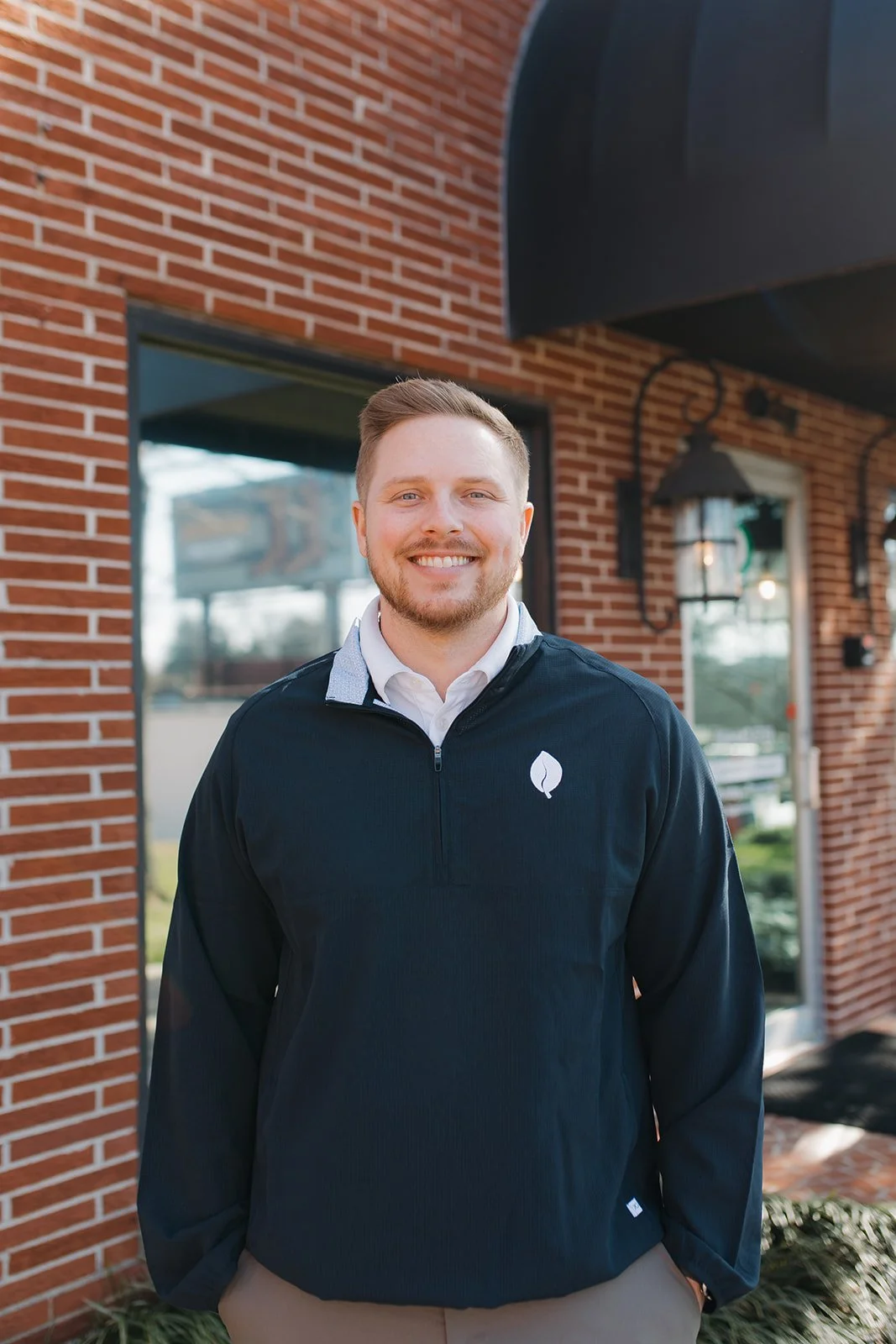 A smiling Chiropractor with light brown hair and beard, wearing a black jacket with a white logo, standing outdoors in front of a brick building.